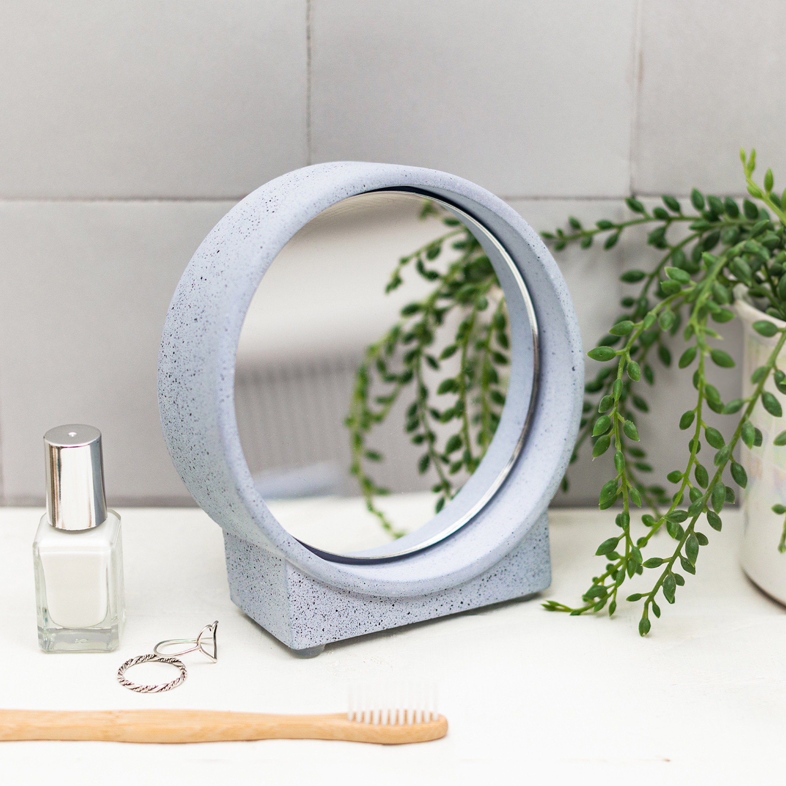 Decorative mirror with lilac speckled frame, white background with green plant, nail polish, jewellery, and wooden toothbrush on a white surface.