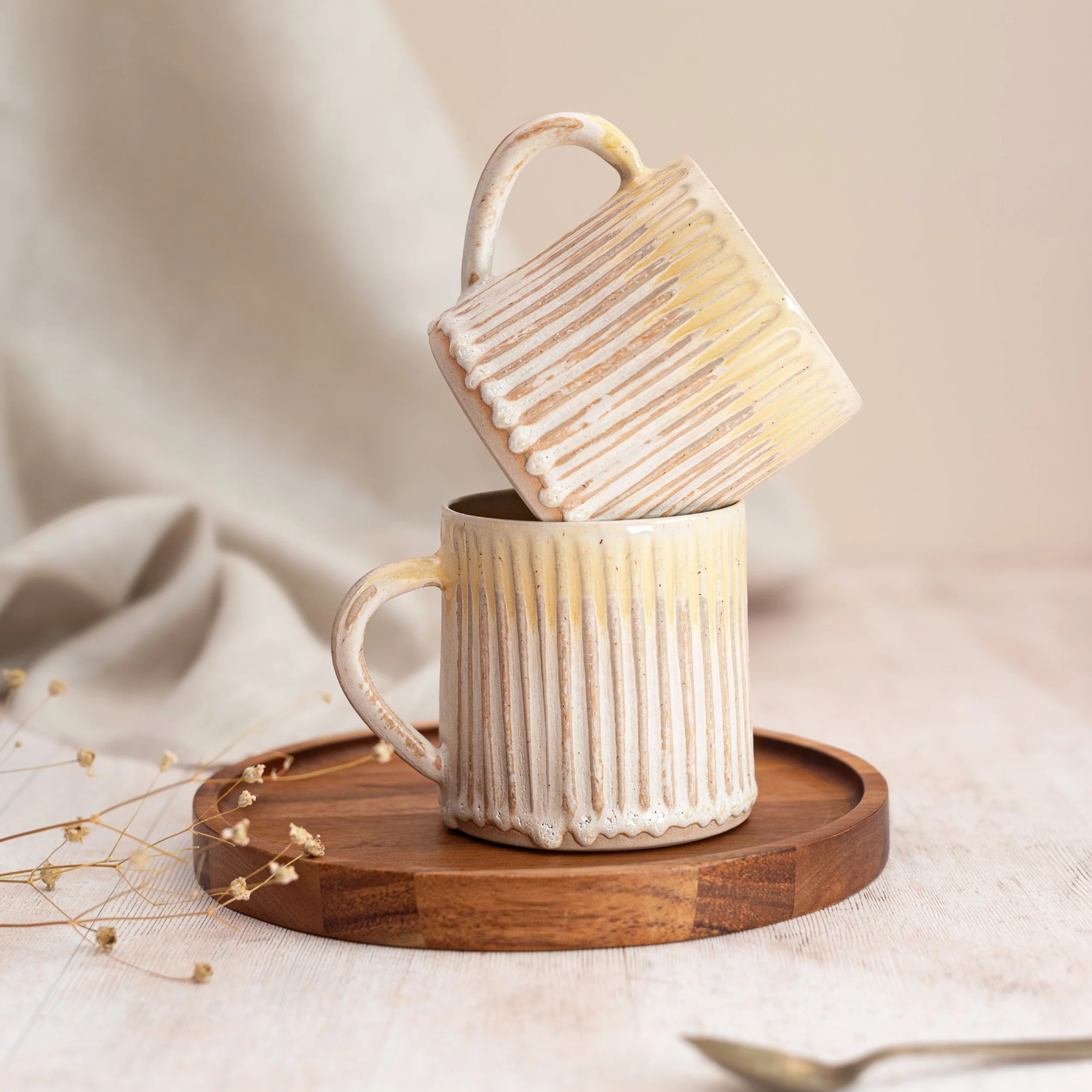 Two cream-coloured ceramic mugs with a ribbed texture, one stacked inside the other, placed on a round wooden tray. The background includes neutral-coloured fabric and dried flowers.