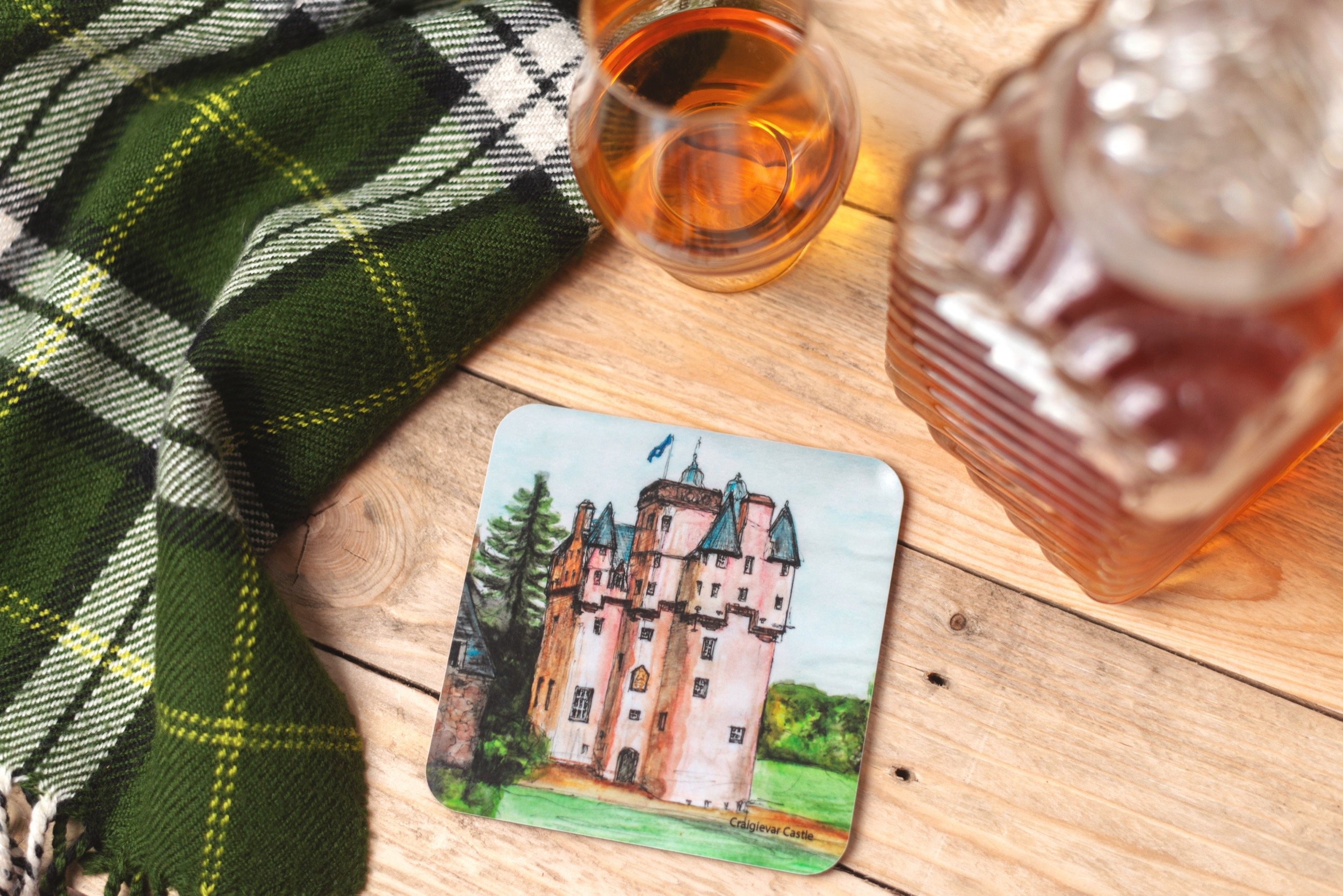 A souvenir coaster featuring a watercolour painting of Craigevar Castle. The coaster is on a wooden surface next to a green plaid fabric, a small glass of whisky, and a larger jar of the same beverage.