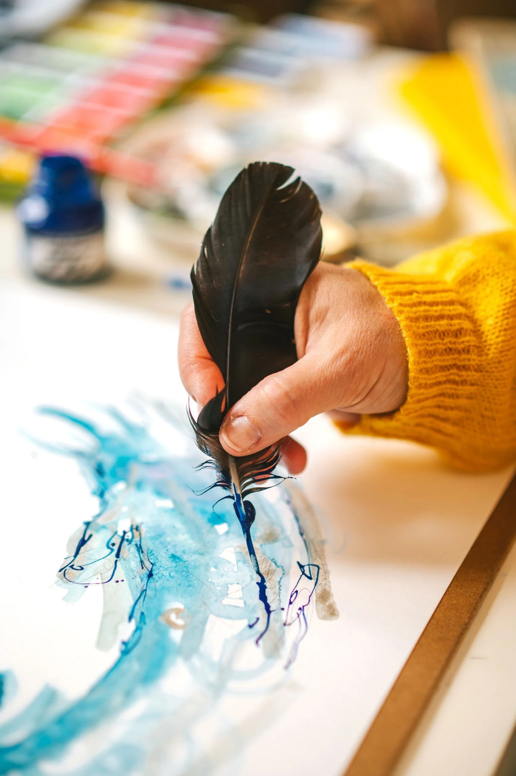 Person holding a black feather quill pen and creating watercolor art on paper, with art supplies in the background.