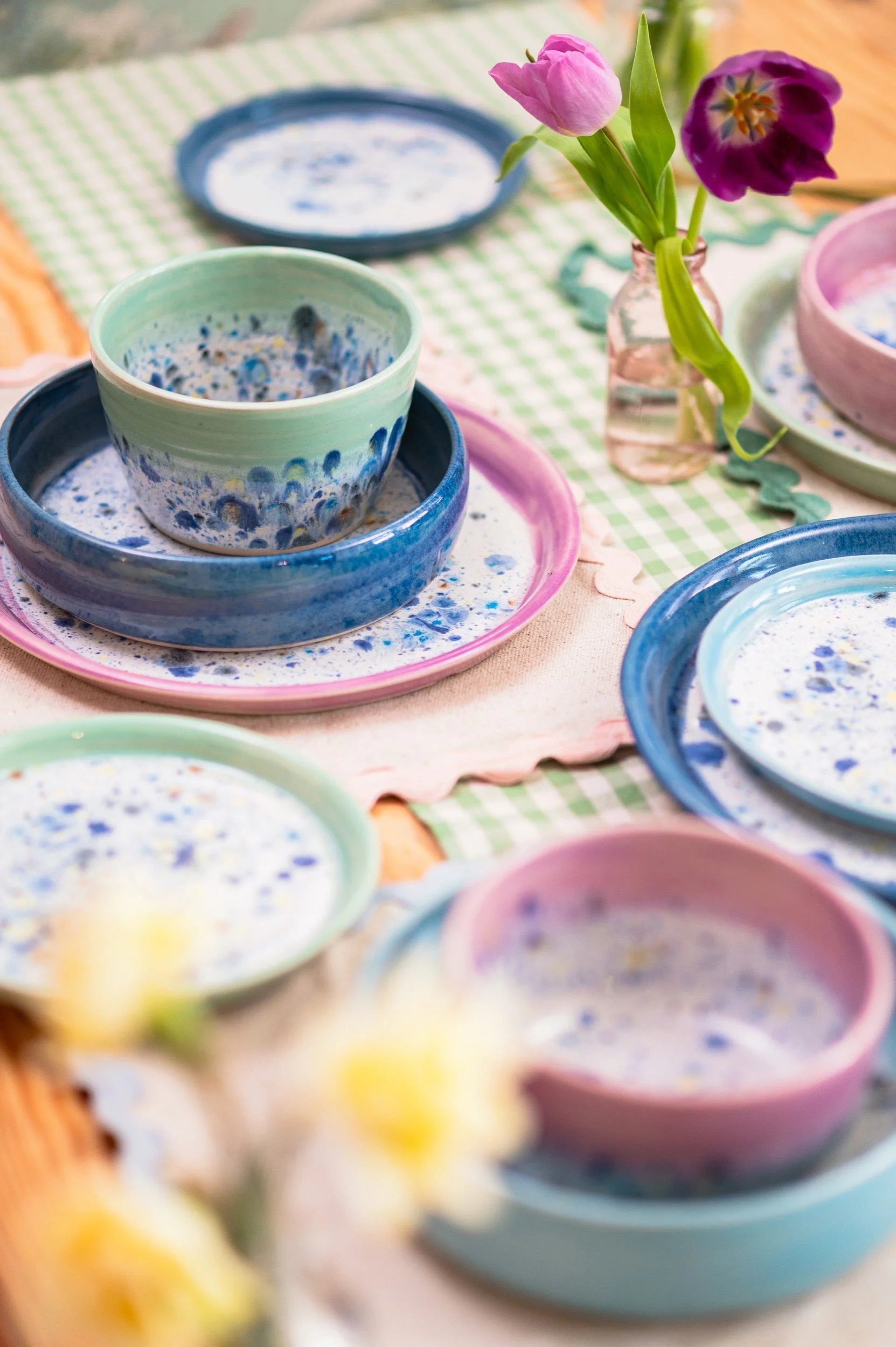 Colorful ceramic bowls and plates with a blue and white speckled pattern, arranged on a table with a pink and a green placemat, and a small glass vase with pink and purple tulips.