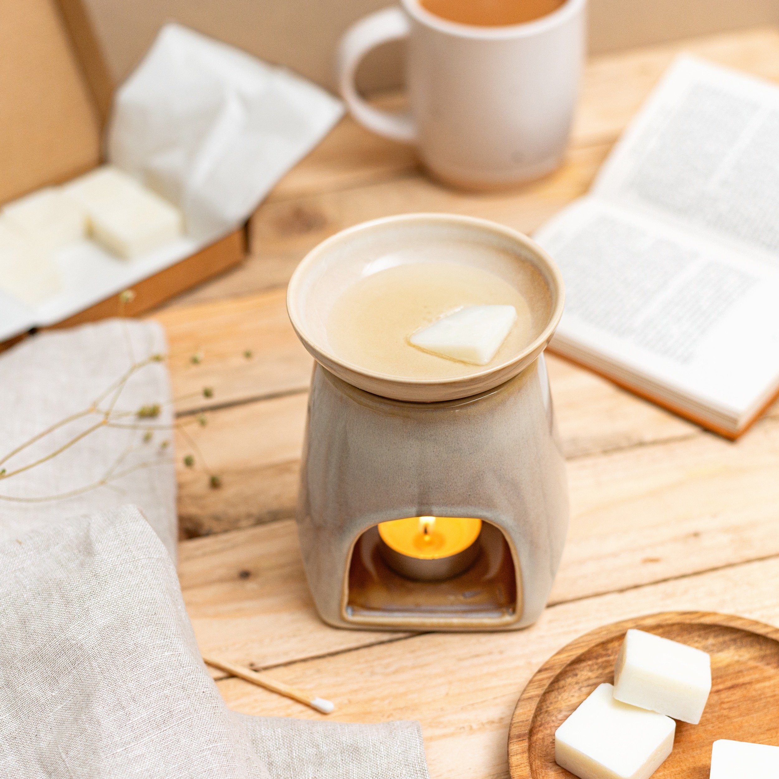 A candle warmer on a wooden table with melting wax, a cup of tea in a white mug, an open book, a wooden tray with soap, and some linen fabric and dried plants.