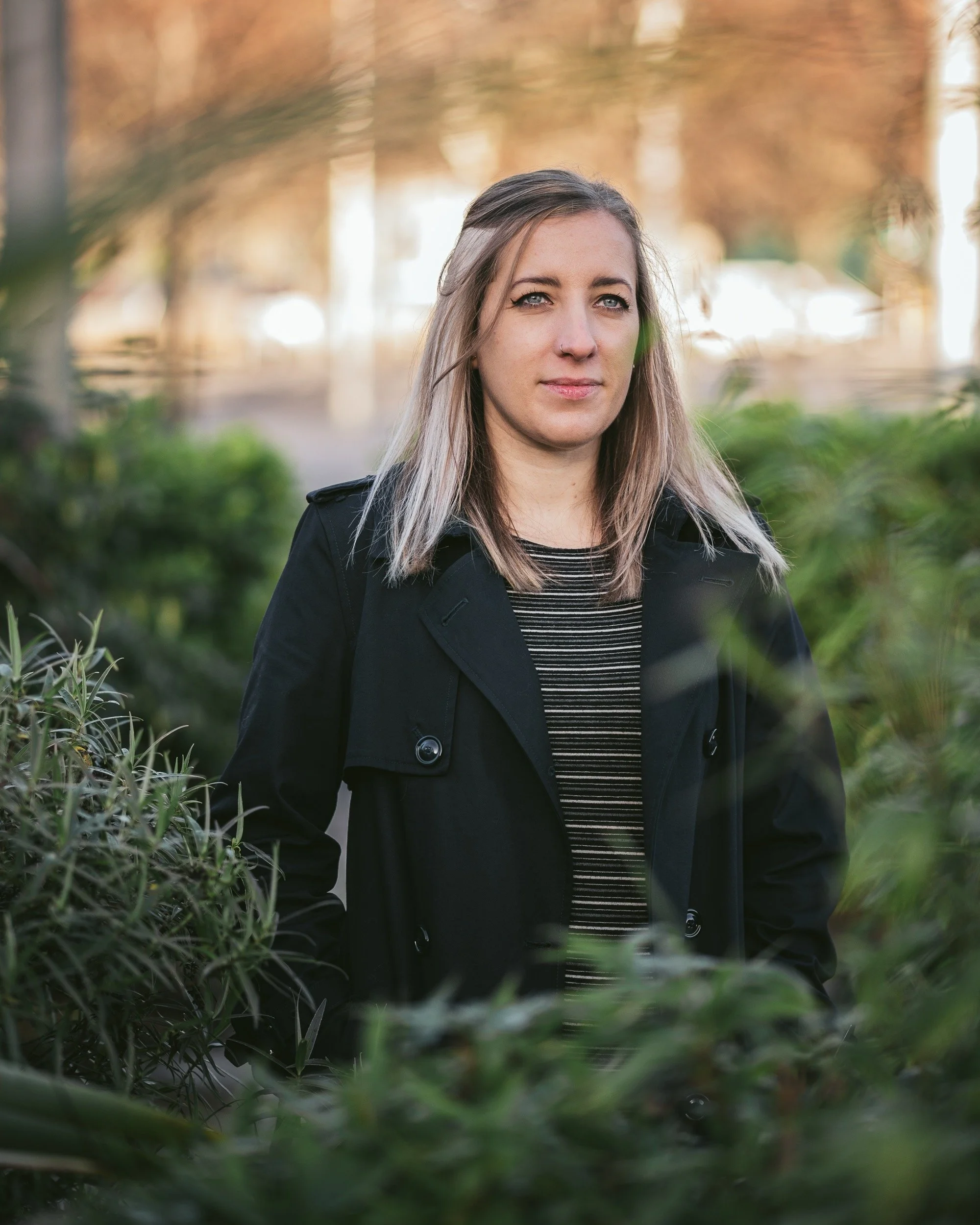 A woman with blonde hair and a nose piercing standing outdoors among green foliage, wearing a black coat and a striped shirt, with a blurred background of trees and sunlight.
