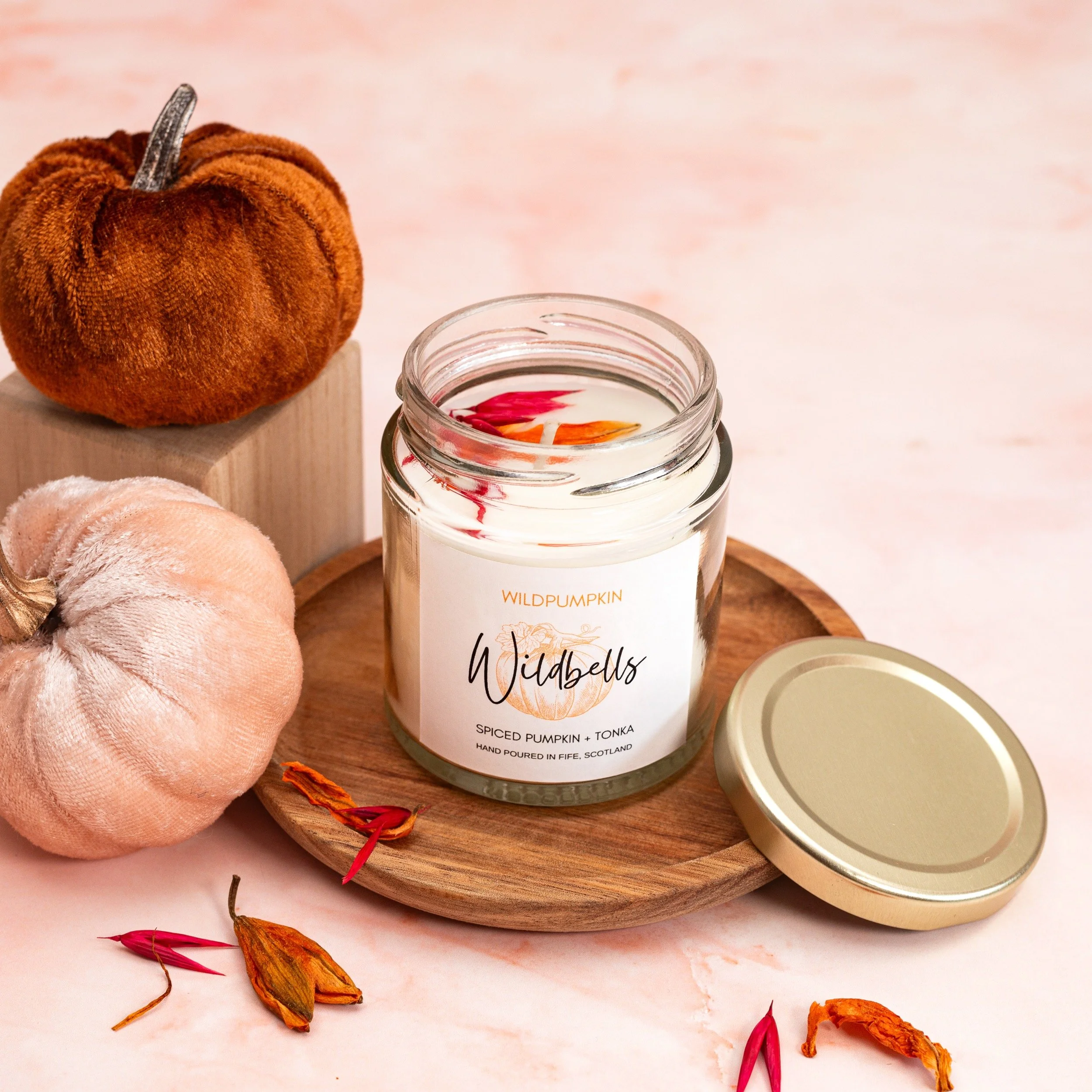 A lit scented candle in a glass jar labeled 'Wildwells Spiced Pumpkin + Tonka' on a round wooden tray with two plush pumpkins, one pink and one orange, nearby and dried flower petals scattered around.