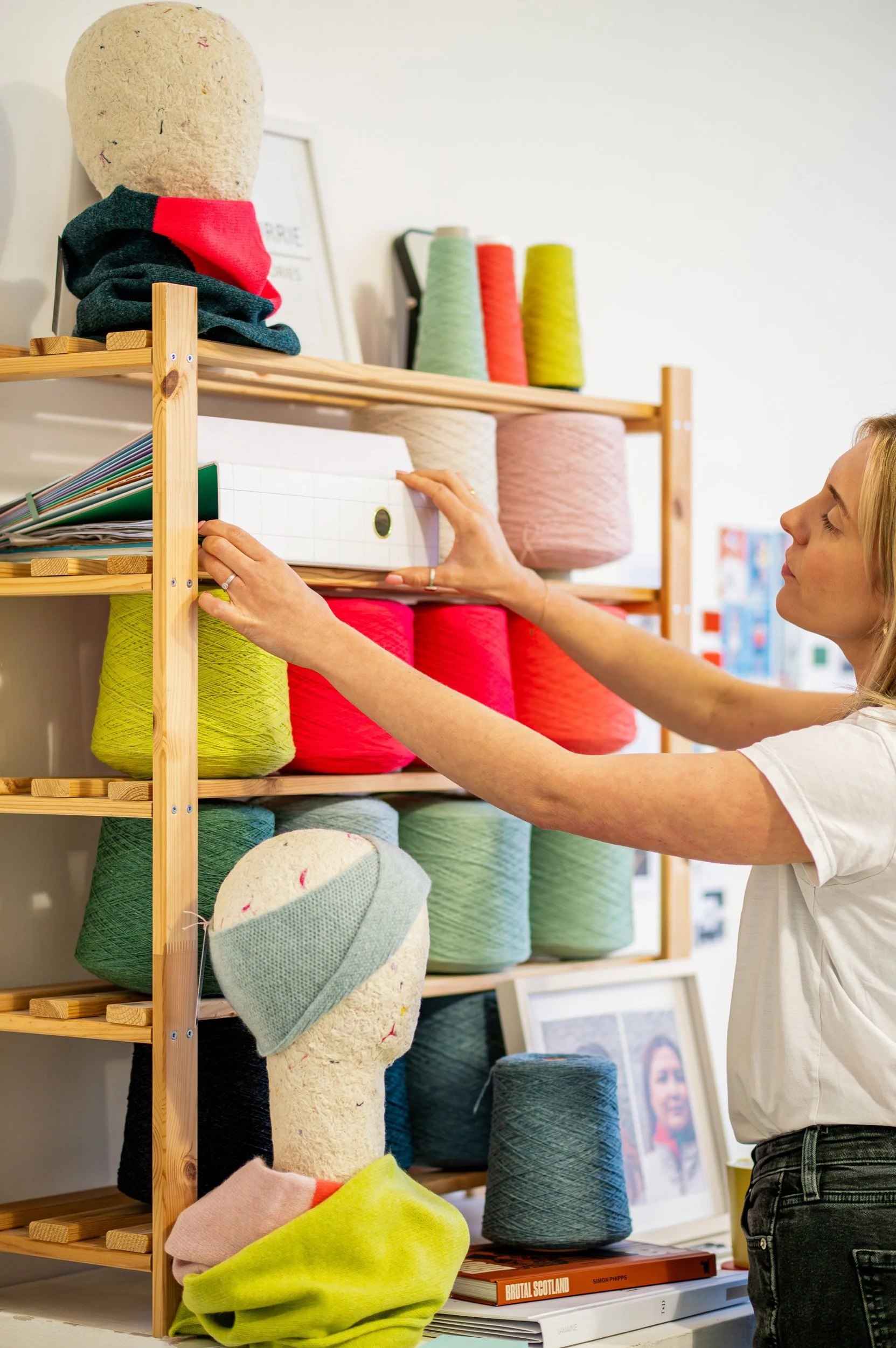 A woman organizing wool and knitting supplies on a wooden shelf in a studio, with spools in various colors.