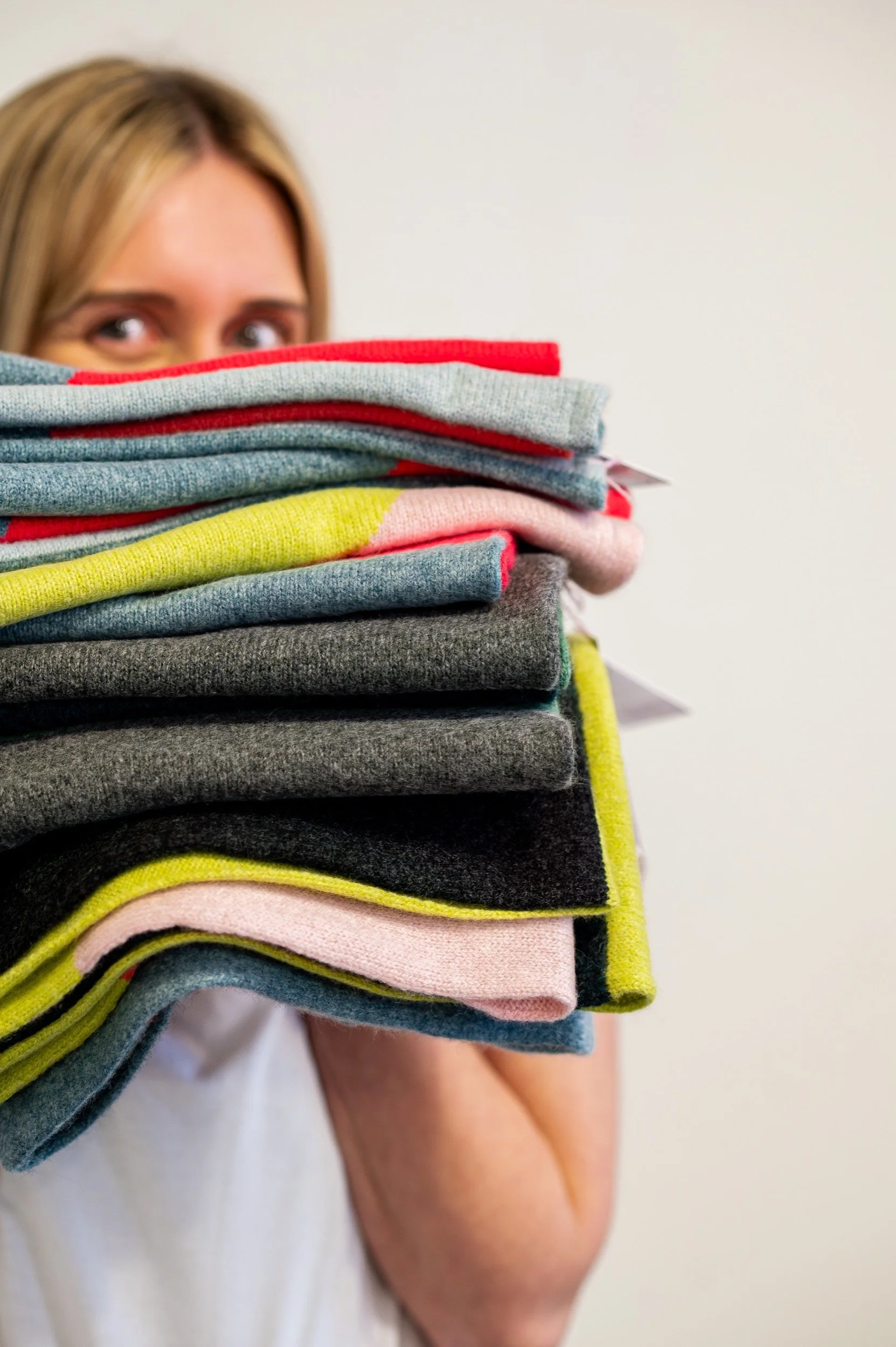 Woman holding a large stack of folded colorful knitted snoods partially hiding her face, with only her eyes and part of her arm visible.