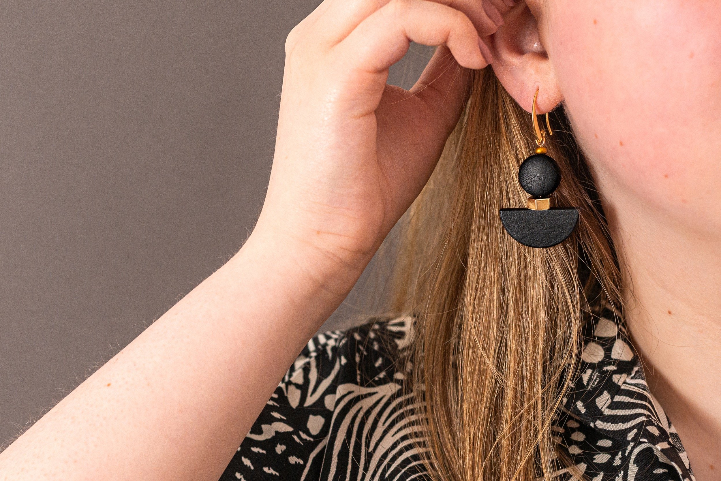 Close-up of a woman wearing a black and white patterned top and a large black geometric earring made of connected semi-circles and a sphere, with her hand near her ear.