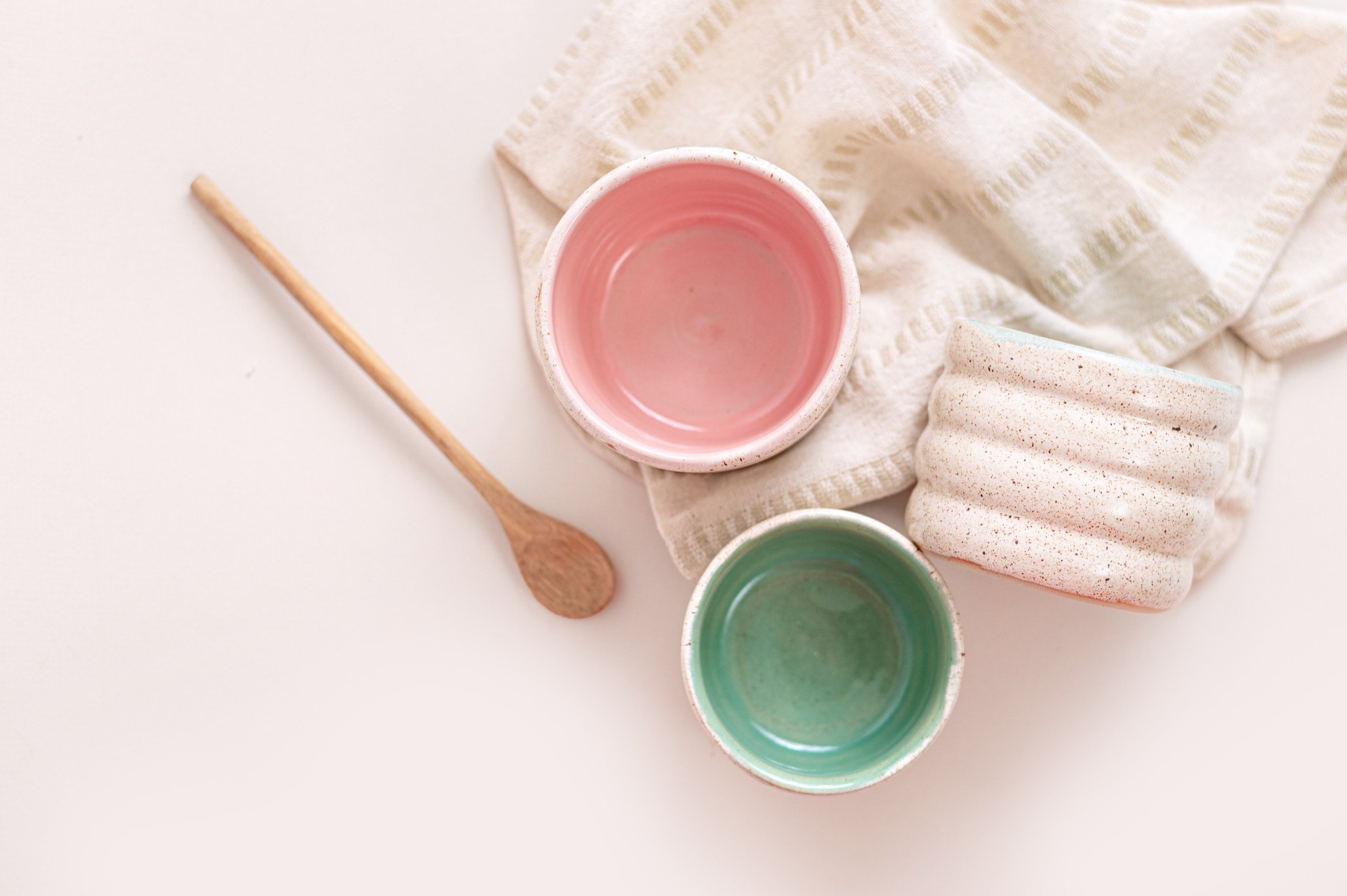 Two small ceramic bowls, one pink and one green, on a white surface with a folded towel and a small wooden spoon nearby.