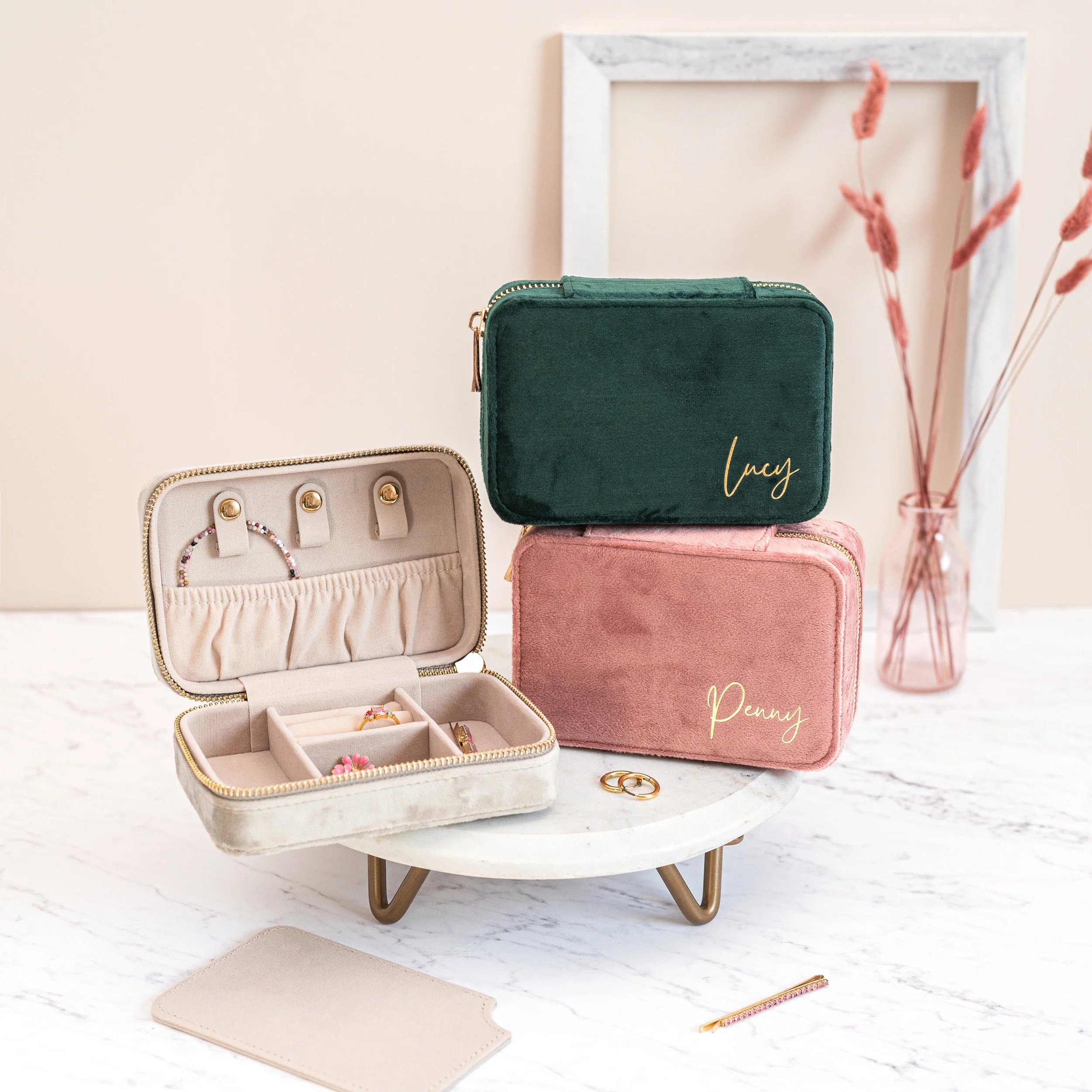Jewellery storage cases including a pink case labeled "Penny," a green case labeled "Lucy," and an open case with jewellery inside. A white marble surface, a framed pink vase with dried pink plants, and gold accessories are also visible.