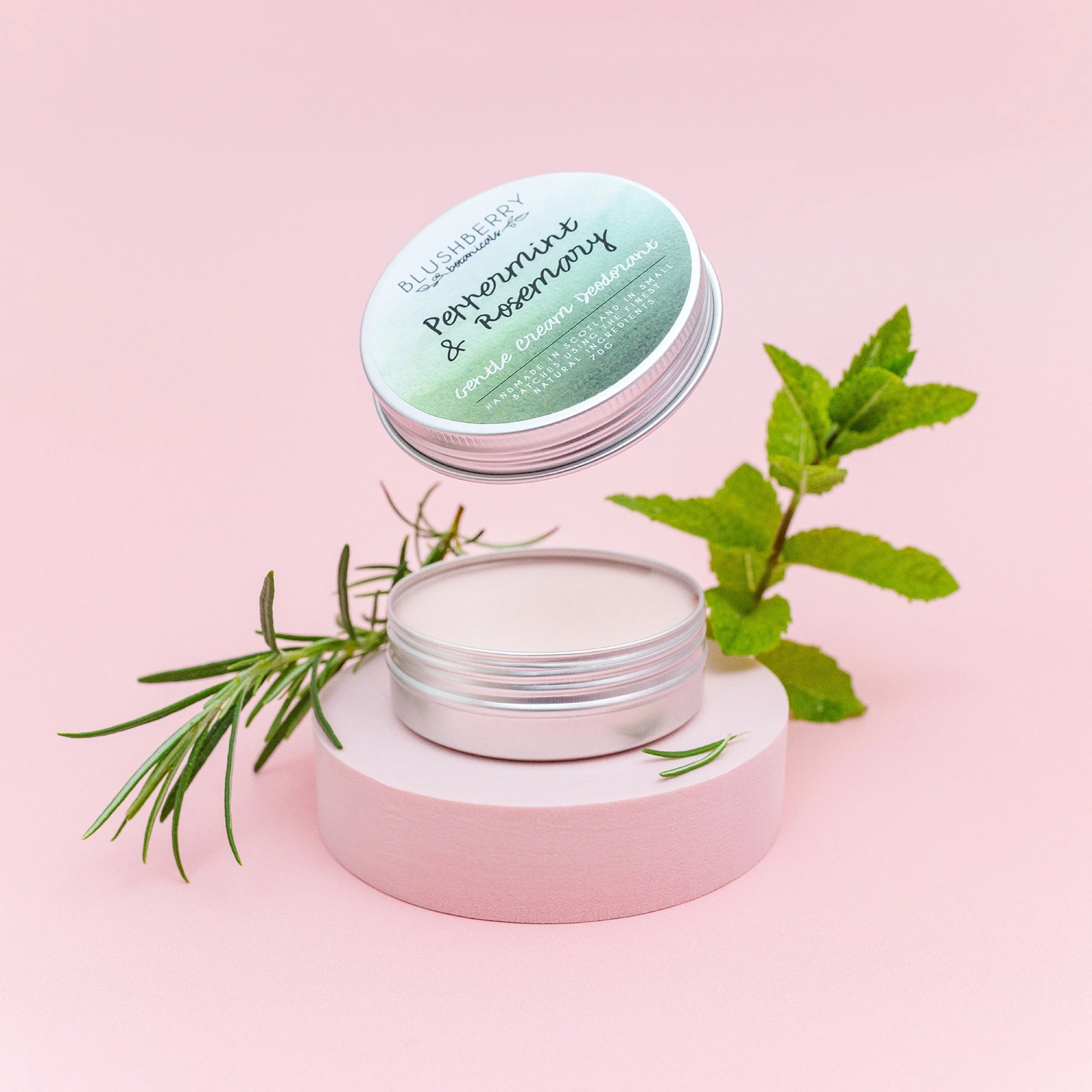 Open tin of cream deodorant with fresh rosemary and mint leaves on pastel pink background.