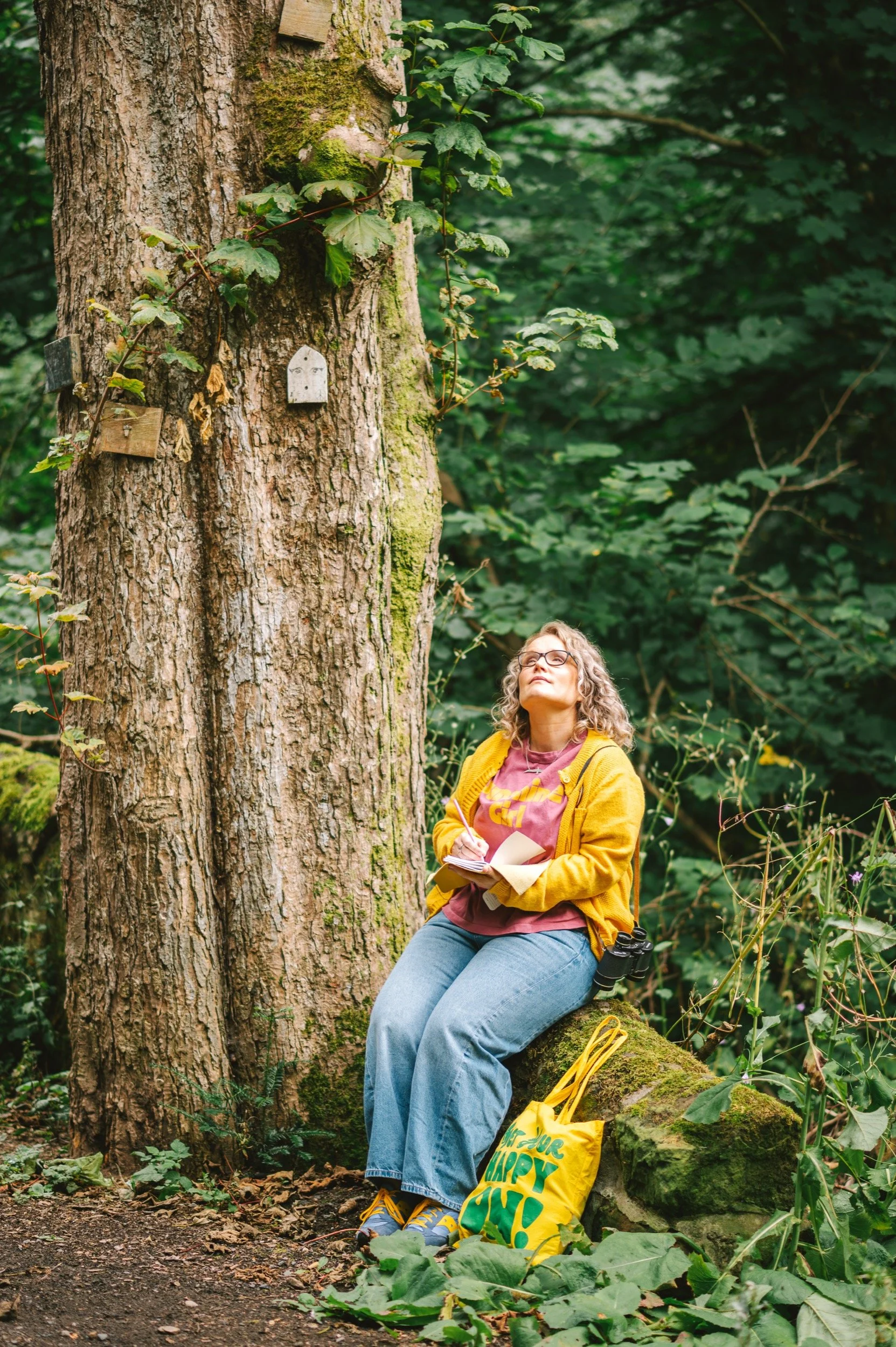 Jenna sits on a wall in a forest, she's holding a sketchbook and looking up into the trees