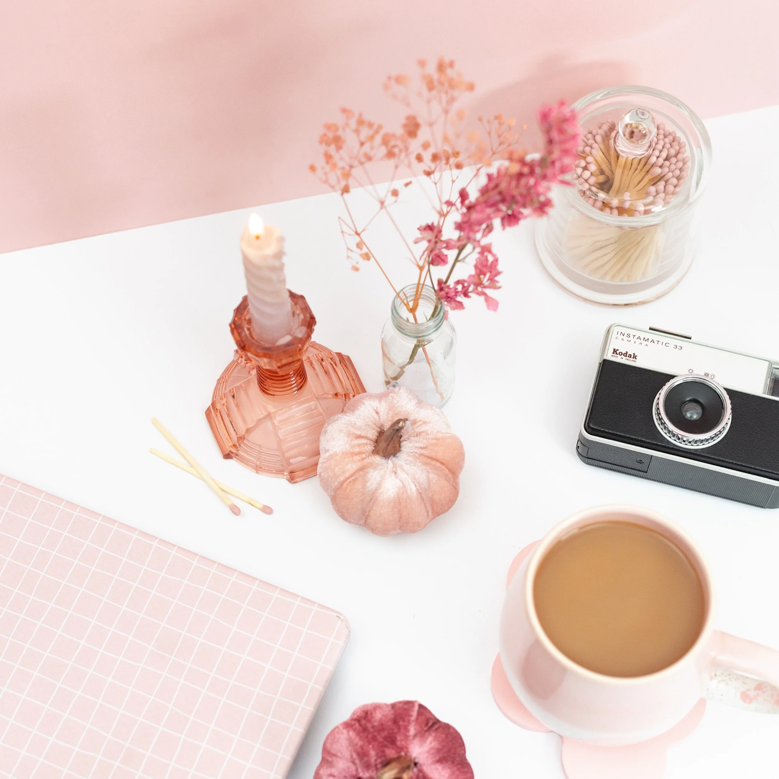 Pink-themed flat lay of decor and personal items, including a lit candle in a pink glass holder, a small pumpkin, a pink mug filled with coffee or tea, a vintage camera, a jar with flowers, a container of pink-tipped matches, and a closed pink notebo