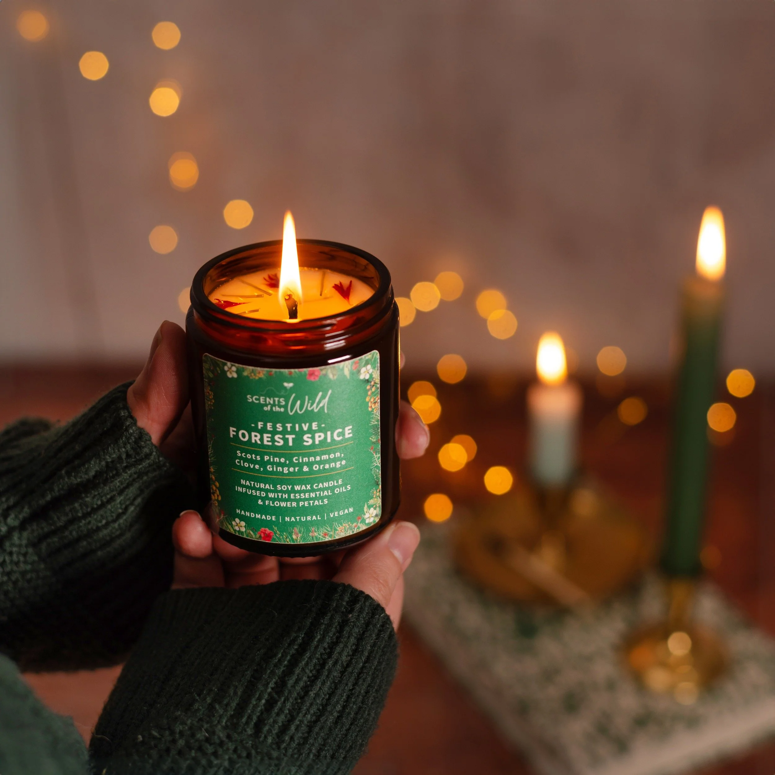 Person holding a lit scented candle labeled 'Festive Forest Spice' with a warm glow and blurred background of other candles and string lights.