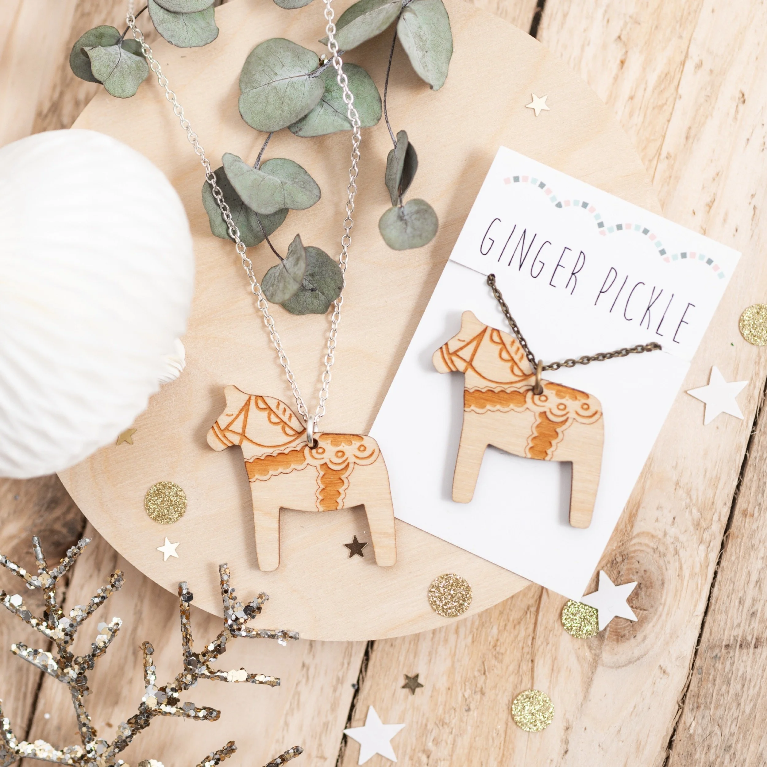 Wooden horse necklaces on display, with eucalyptus leaves, star-shaped and glittery gold confetti around, on a light wooden surface, labeled with a card reading "Ginger Pickle".