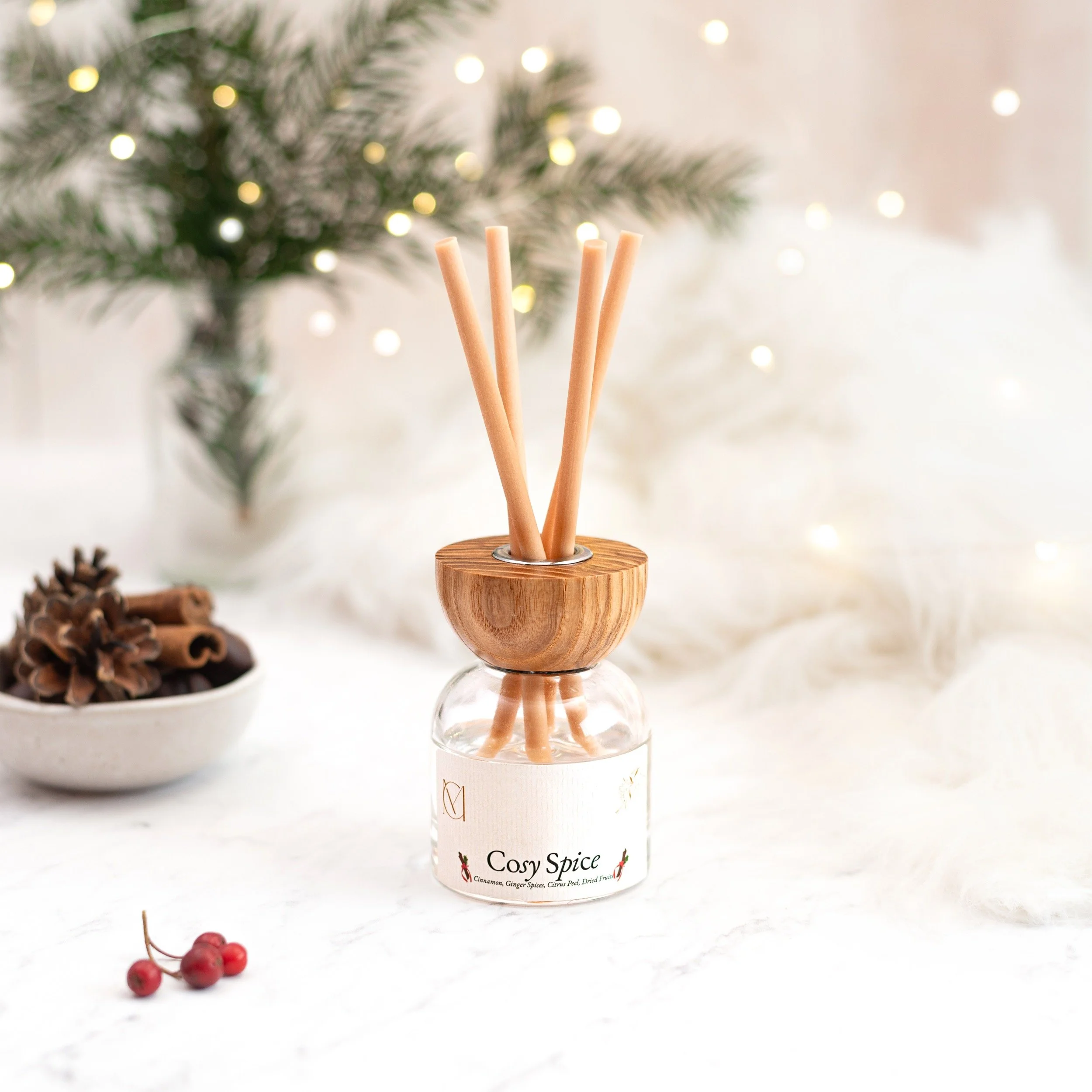 A diffuser with wooden sticks labeled 'Cosy Spice' on a white surface. In the background, there are pinecones, red berries, and blurred Christmas lights and a tree.