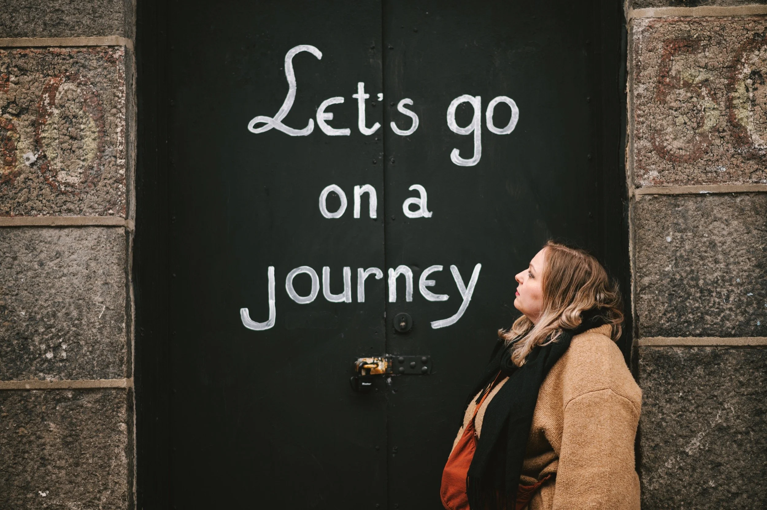 A woman with blonde hair wearing a brown coat and black scarf stands in profile next to a black door with white painted lettering that says, "Let's go on a journey."