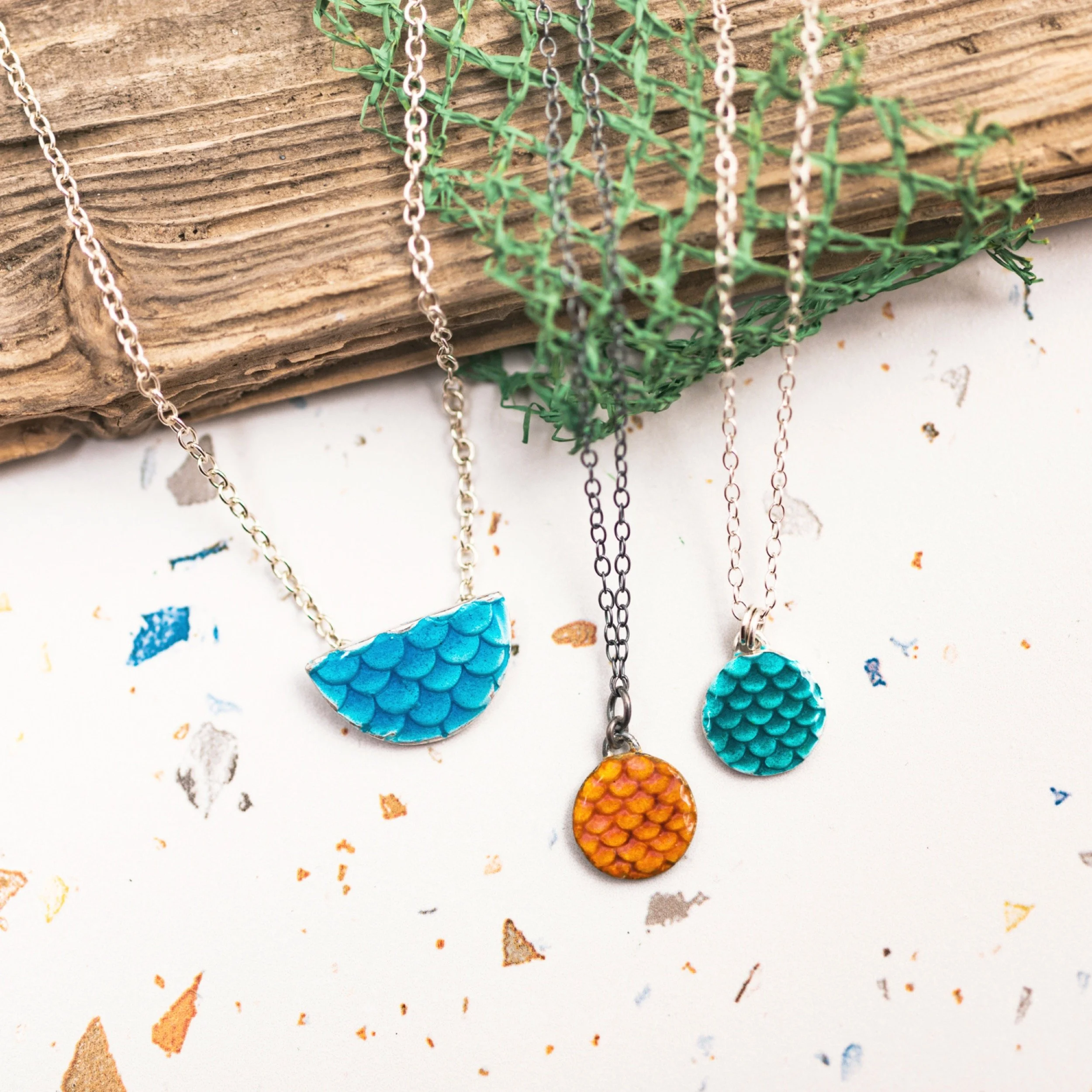 Three necklaces with mermaid scale designs in different colors, hanging on a wooden board and green mesh.