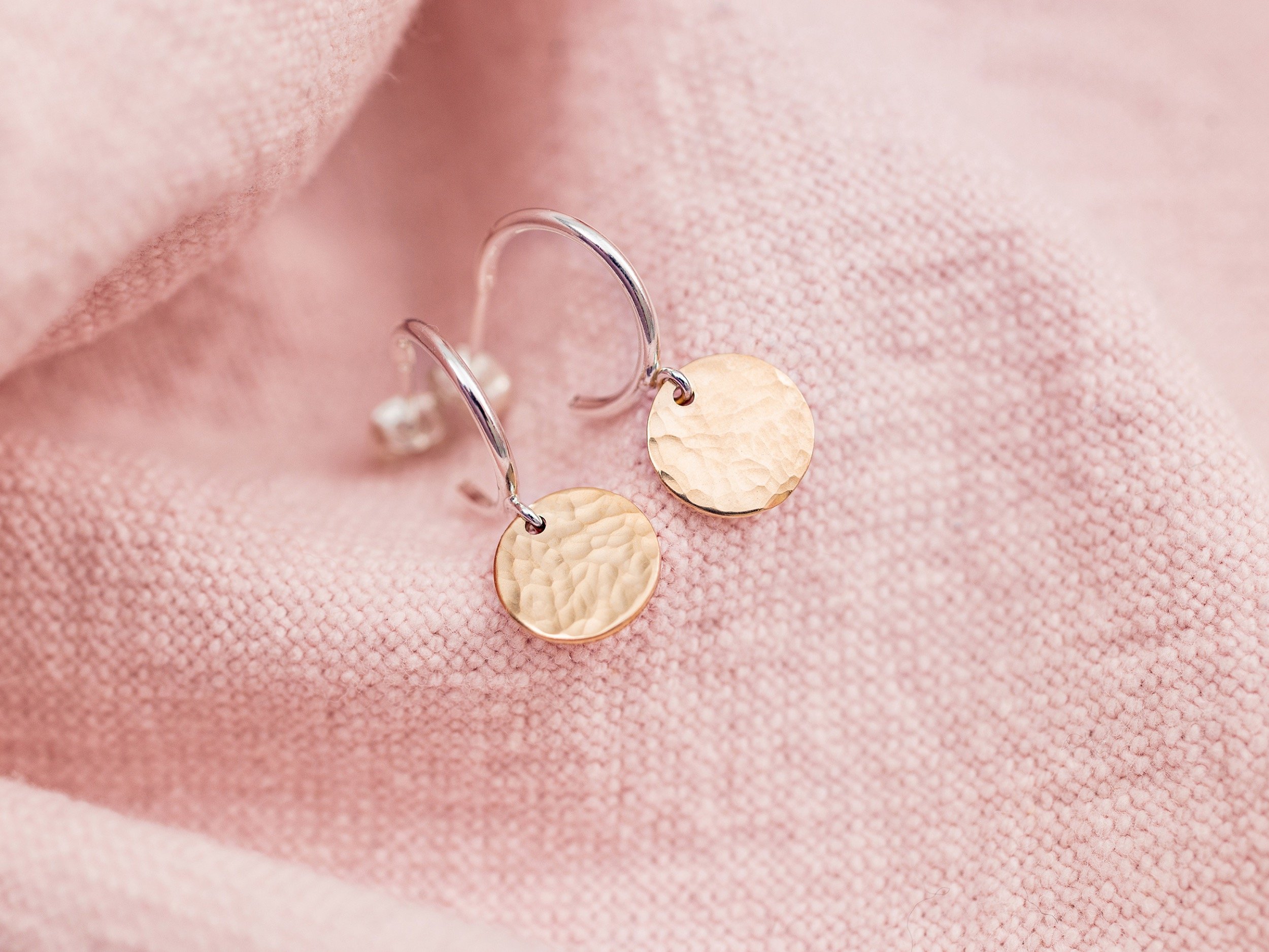 A close-up of gold hammered disc earrings on soft pink textured fabric.