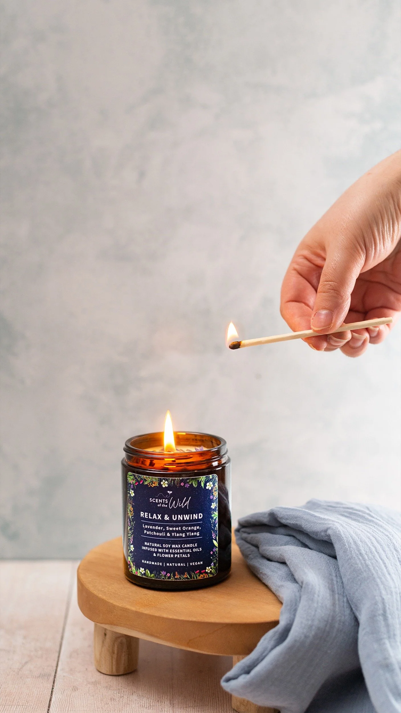 A hand lighting a candle with a matchstick next to a lit candle in a jar labeled 'Scents of the Wild' on a small wooden table, with a blue cloth nearby.