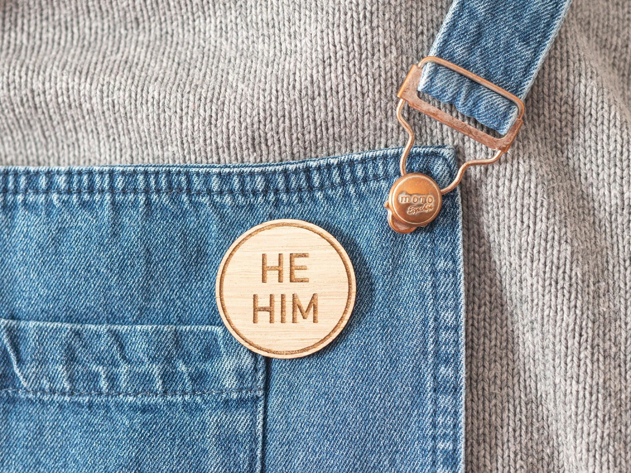 Close-up of a person wearing a gray sweater and blue denim overalls with a wooden badge reading 'HE HIM' and a copper-colored action figure toy attached to the strap.