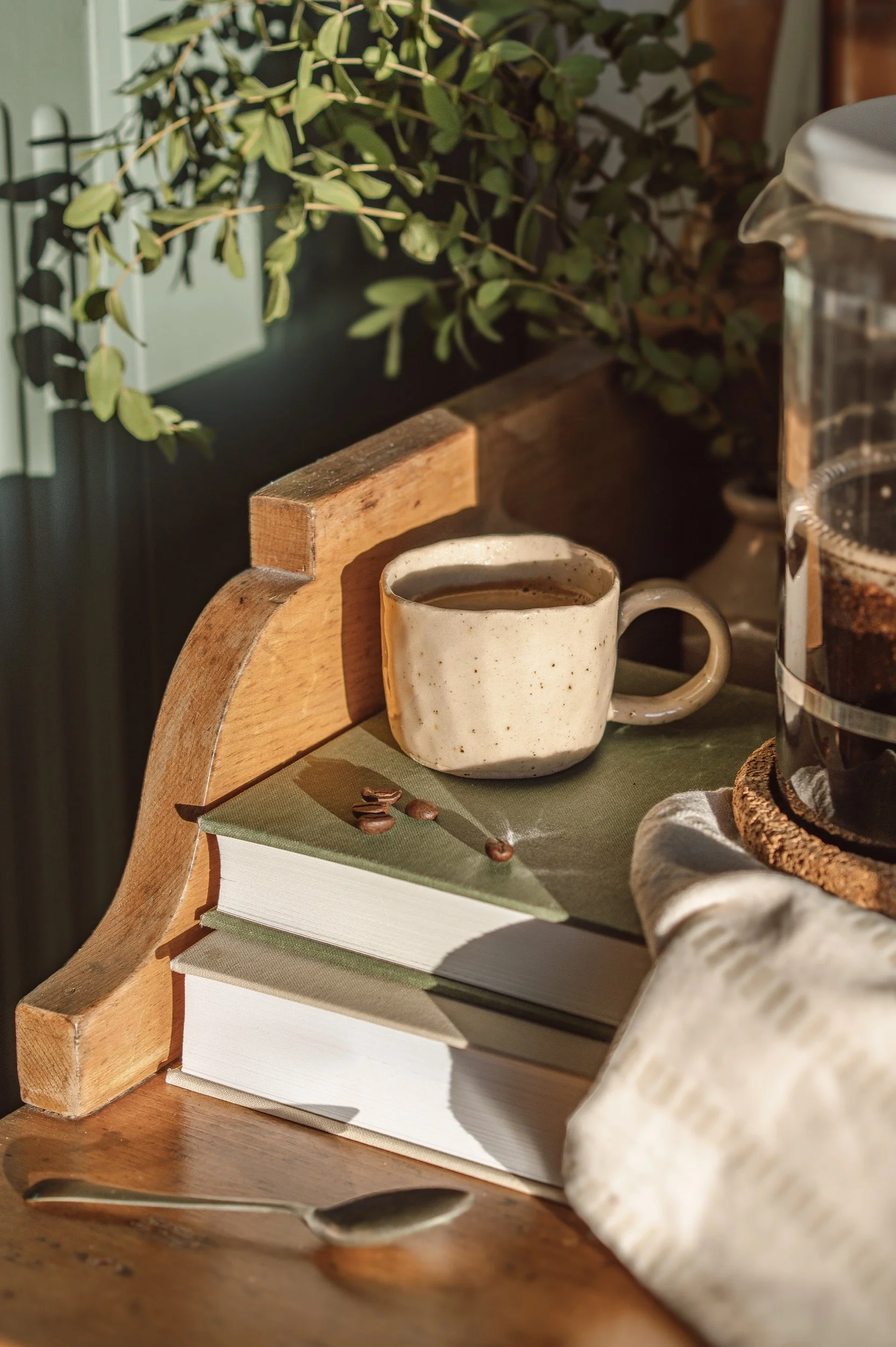 A rustic scene featuring a ceramic coffee mug on top of two stacked books, with a small pile of coffee beans, beside a French press filled with brewed coffee. In the background, there are green leafy plants.
