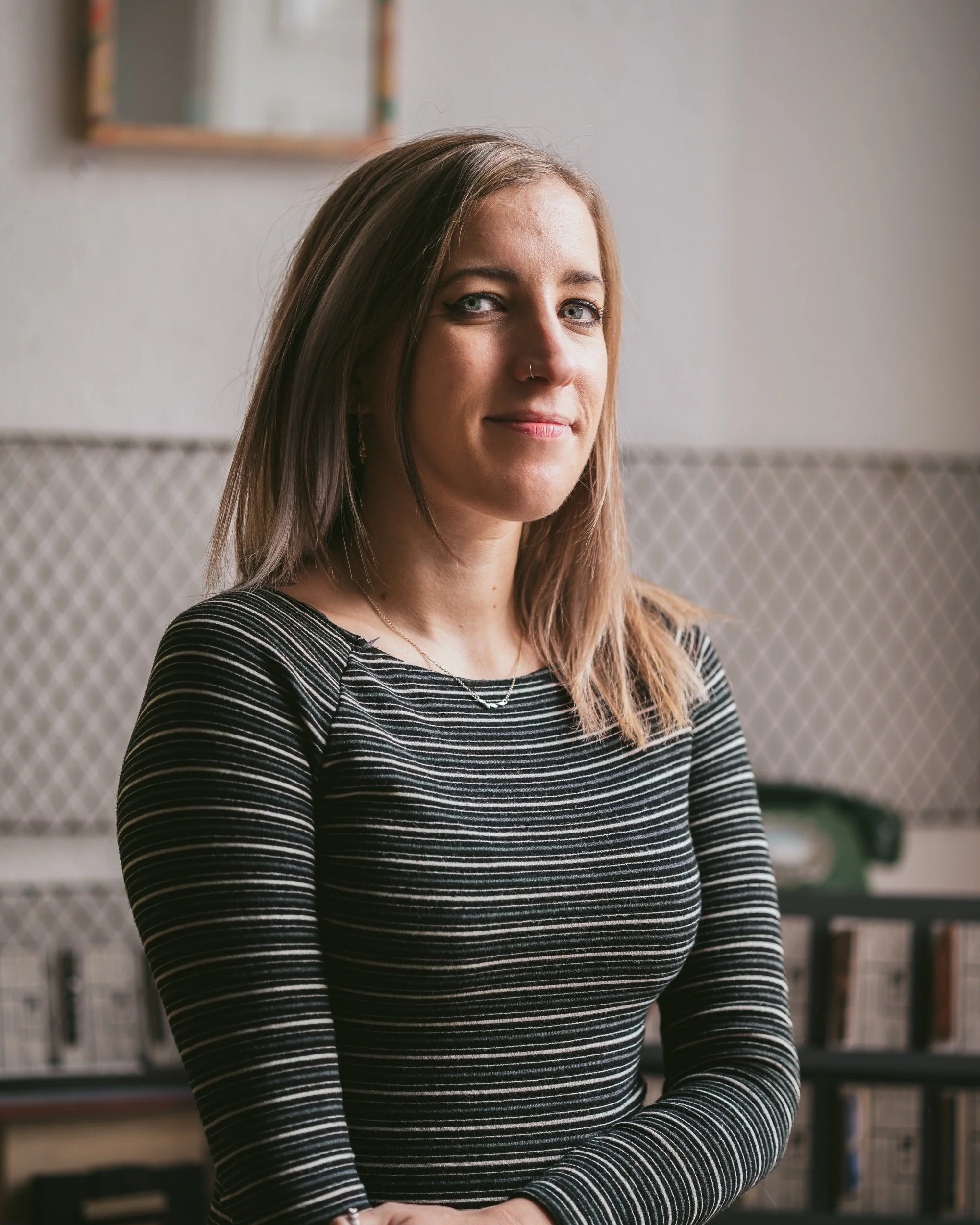 A young woman with shoulder-length light brown hair, fair skin, and blue eyes, wearing a black and white striped long-sleeve shirt, standing indoors with a neutral background of bookshelves and a wall.