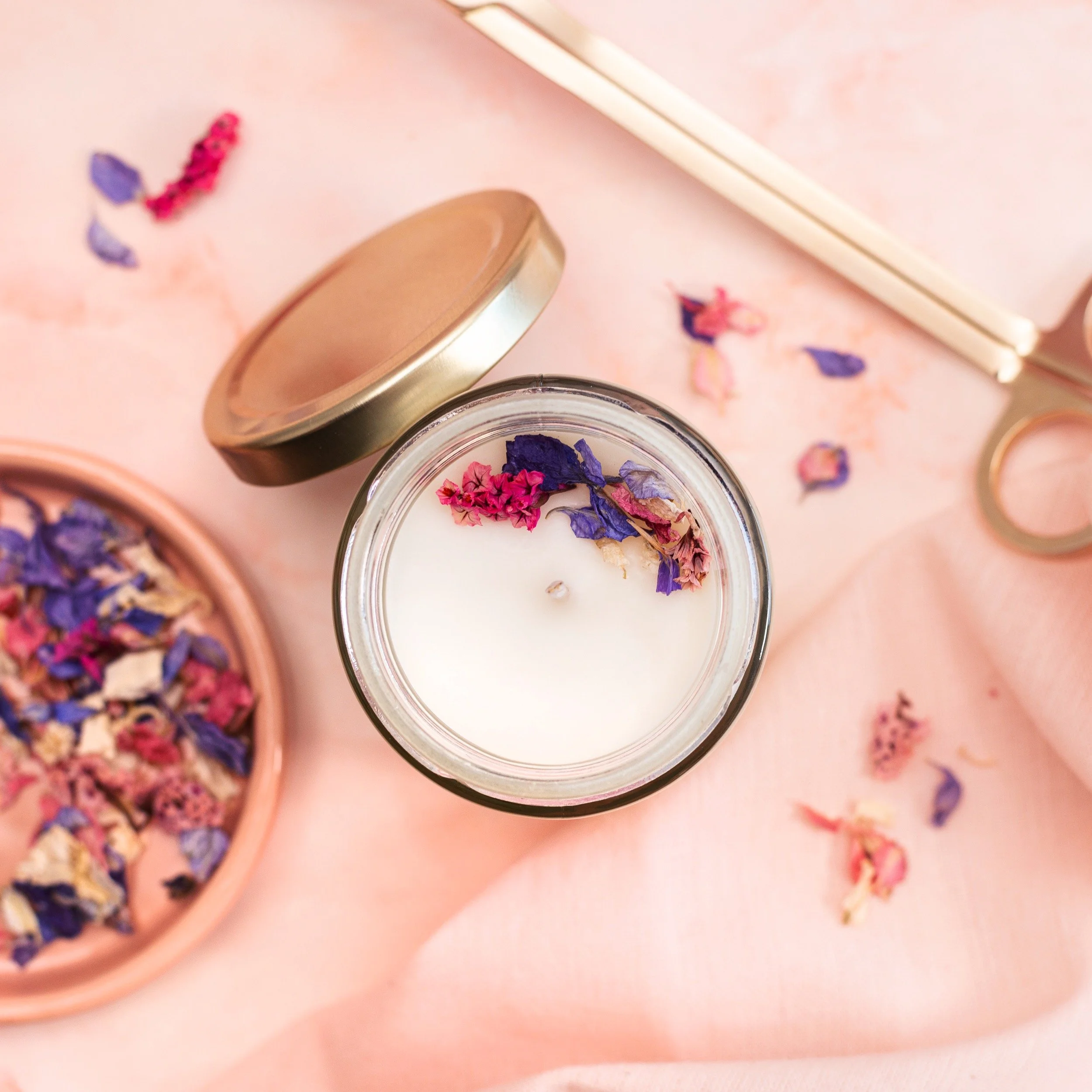 A white candle with dried pink and purple flower petals on top, surrounded by scattered petals, a pair of white chopsticks, a pink dish with dried flower petals, and a scissors on a pink fabric surface.
