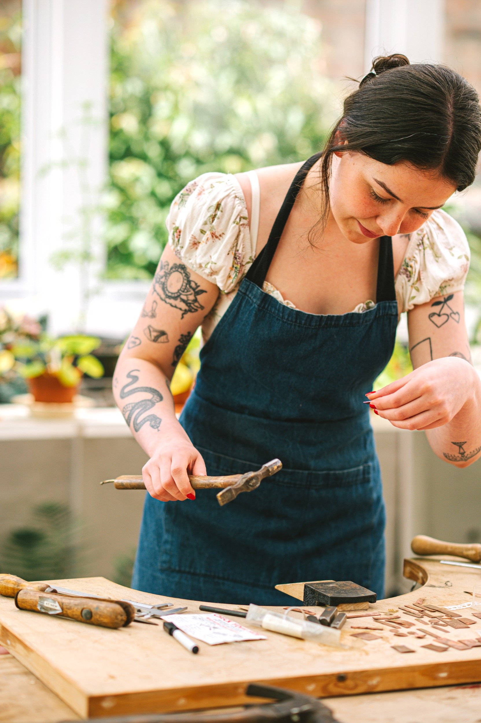 A woman with dark hair and tattoos on her arms working in a workshop,making jewellery. She is wearing a floral blouse and a denim apron, with tools and wooden pieces on the table.