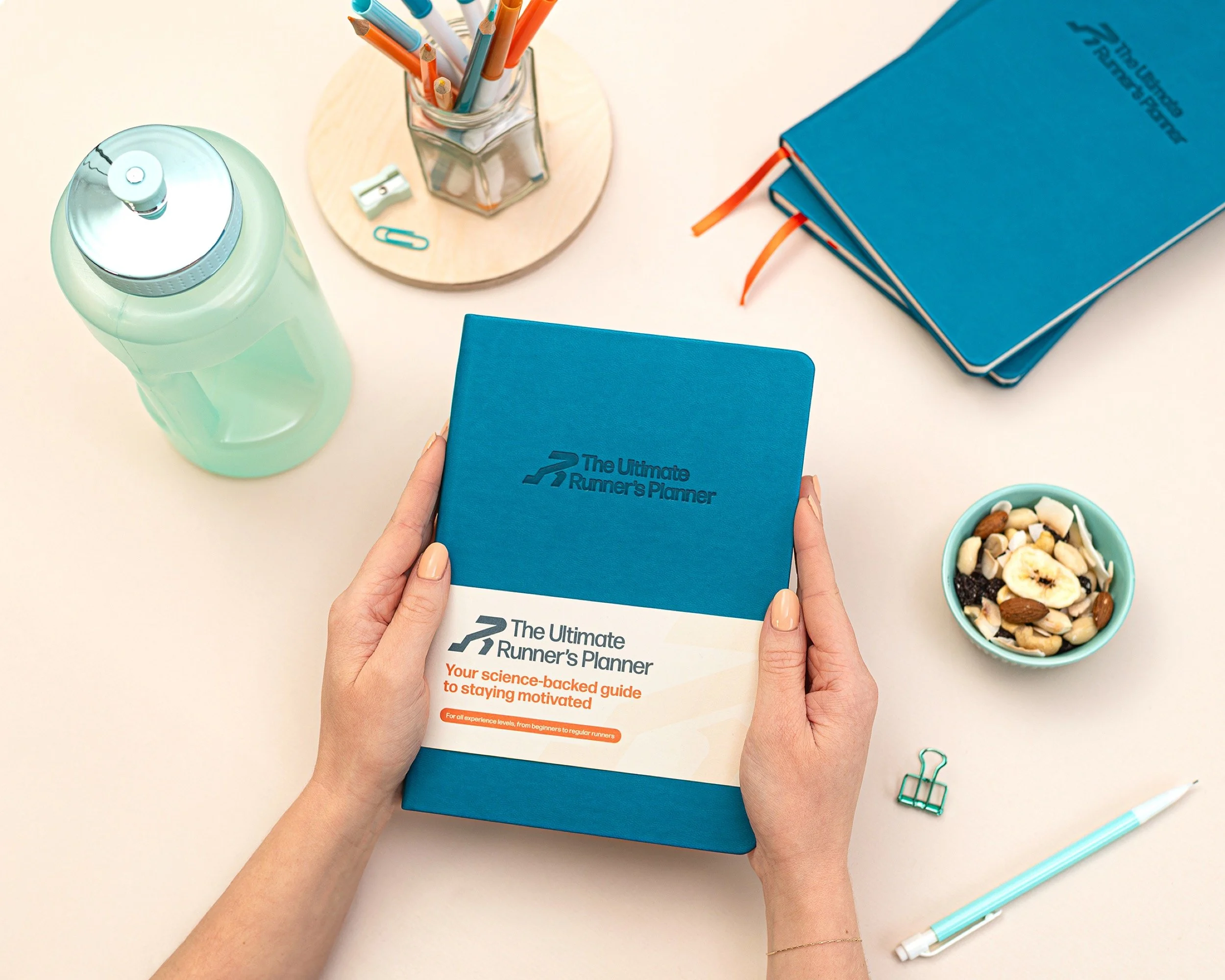 Person holding a blue running planner titled 'The Ultimate Runner's Planner' on a desk, surrounded by a water bottle, pens in a hexagonal glass container, a bowl of trail mix, a stack of blue notebooks, a green binder clip, and a white pen with a tur
