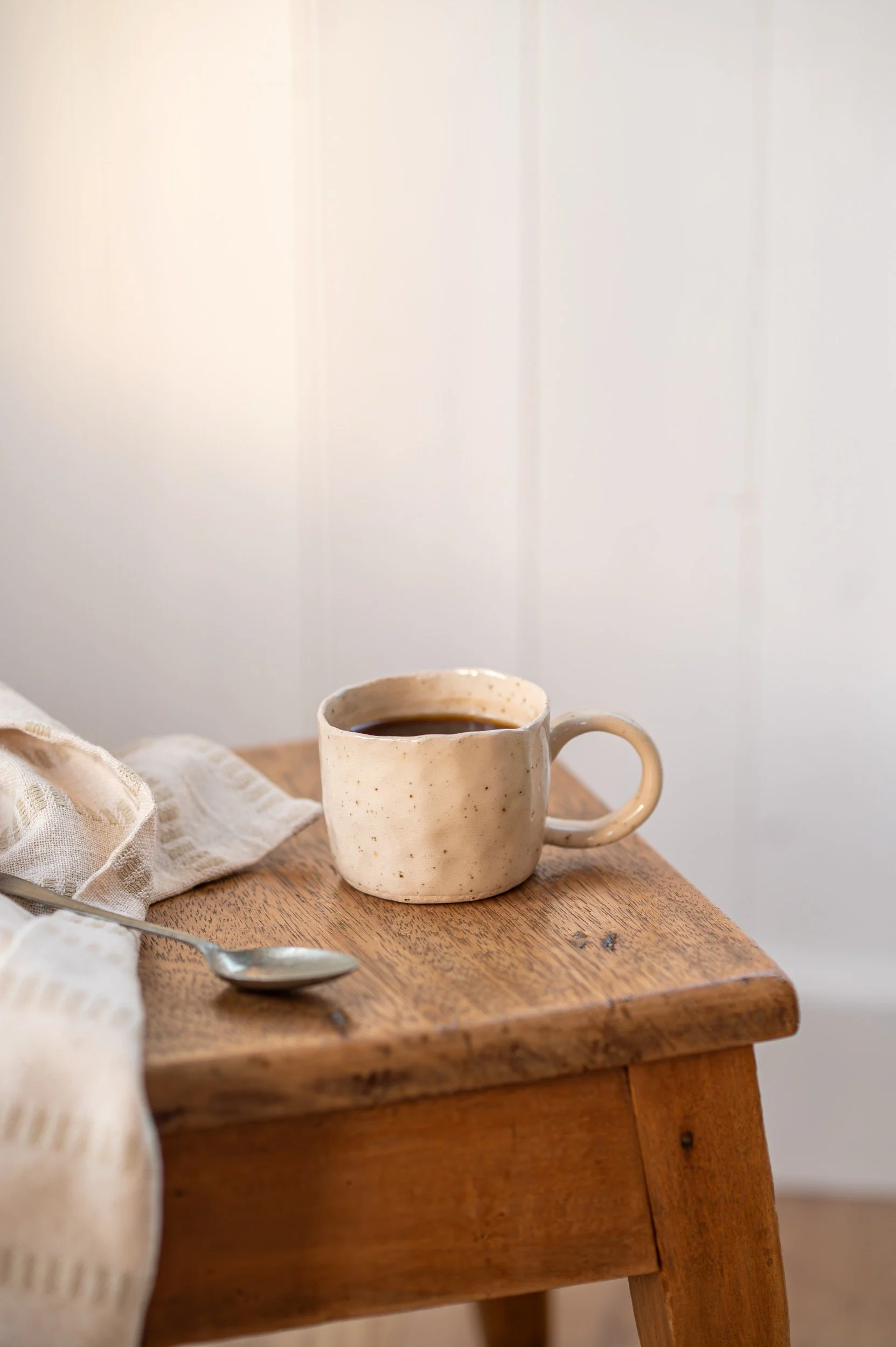A ceramic mug filled with coffee, sitting on a wooden table next to a spoon and a beige cloth.