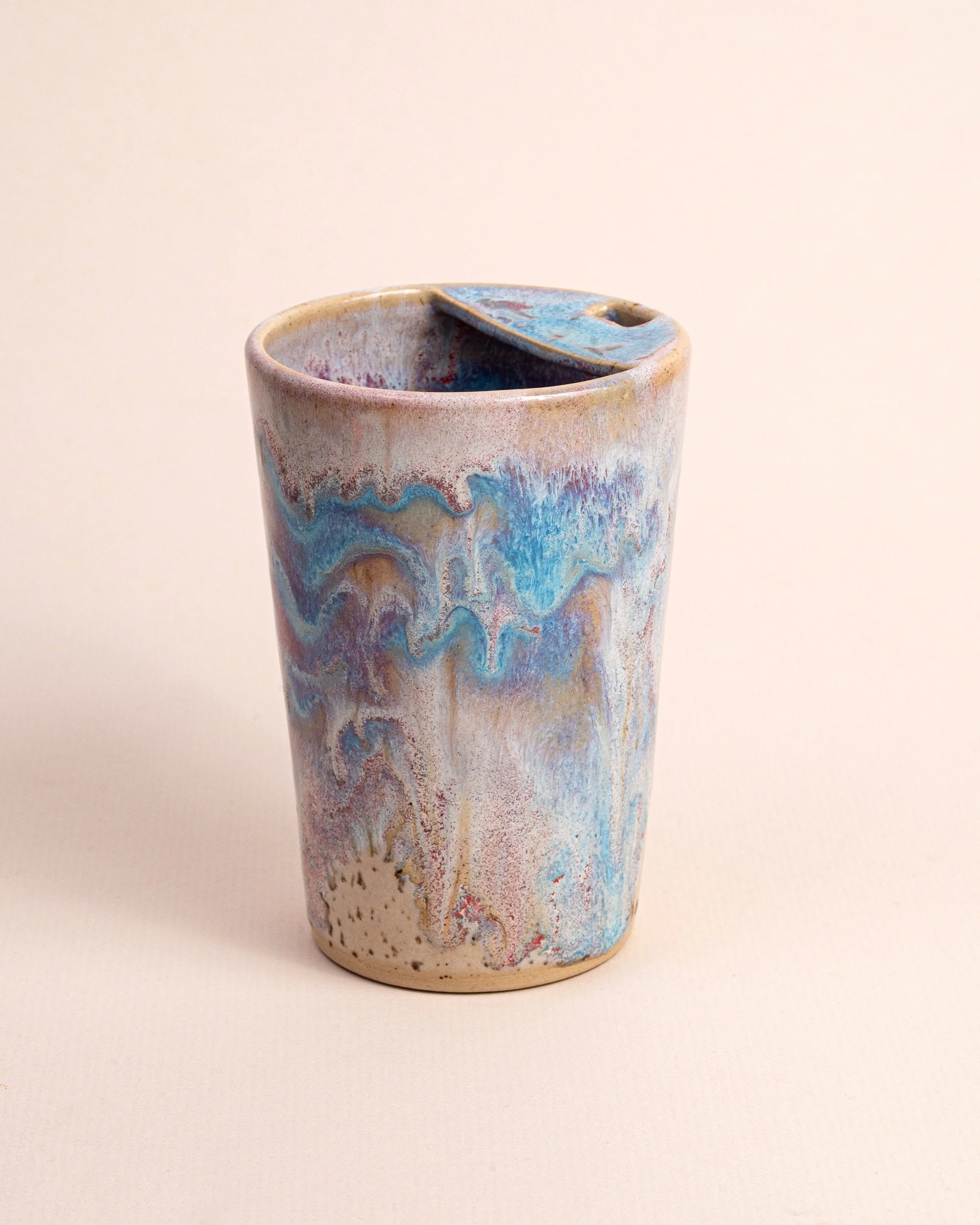 A ceramic mug with a pastel watercolour-like glaze featuring swirling blue, pink, and beige patterns, and a partially open lid.