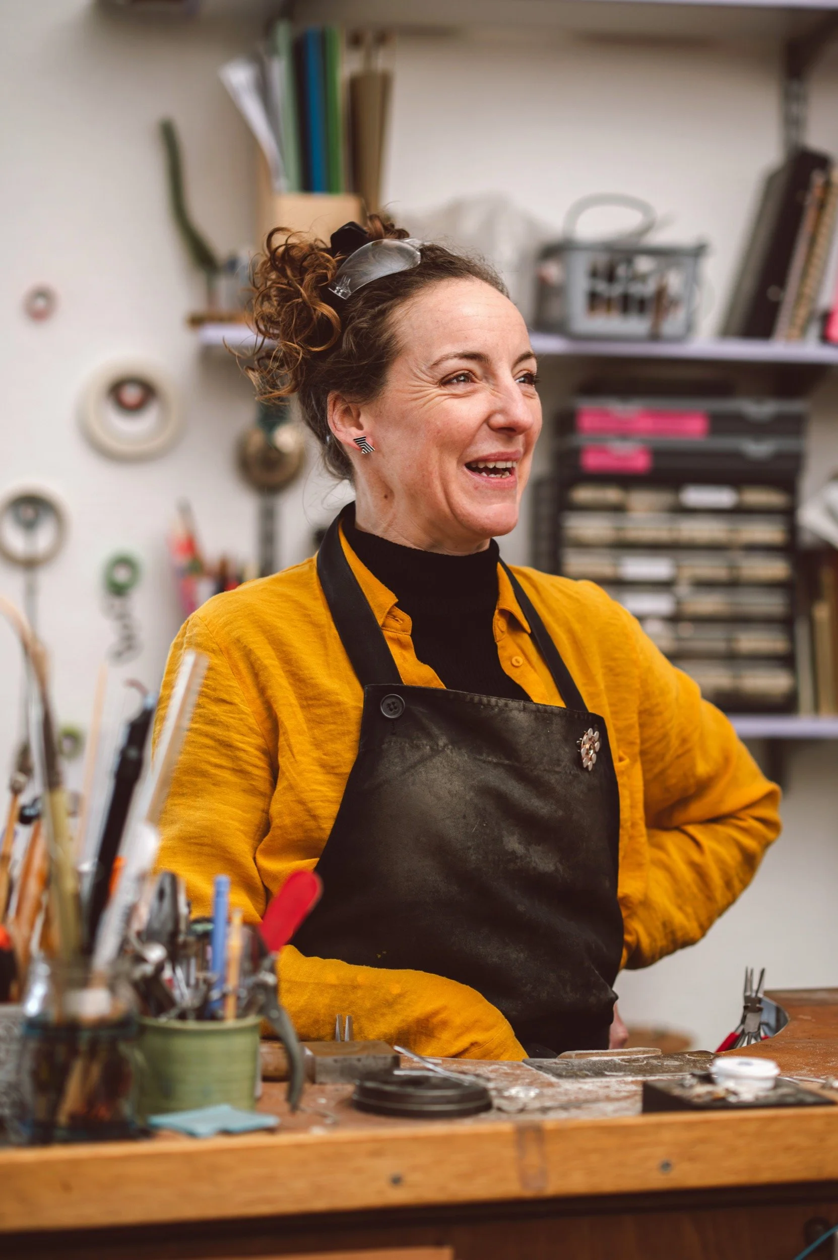 A woman with curly hair tied up wearing a yellow shirt and black apron, smiling in a workshop with various tools and materials in the background.