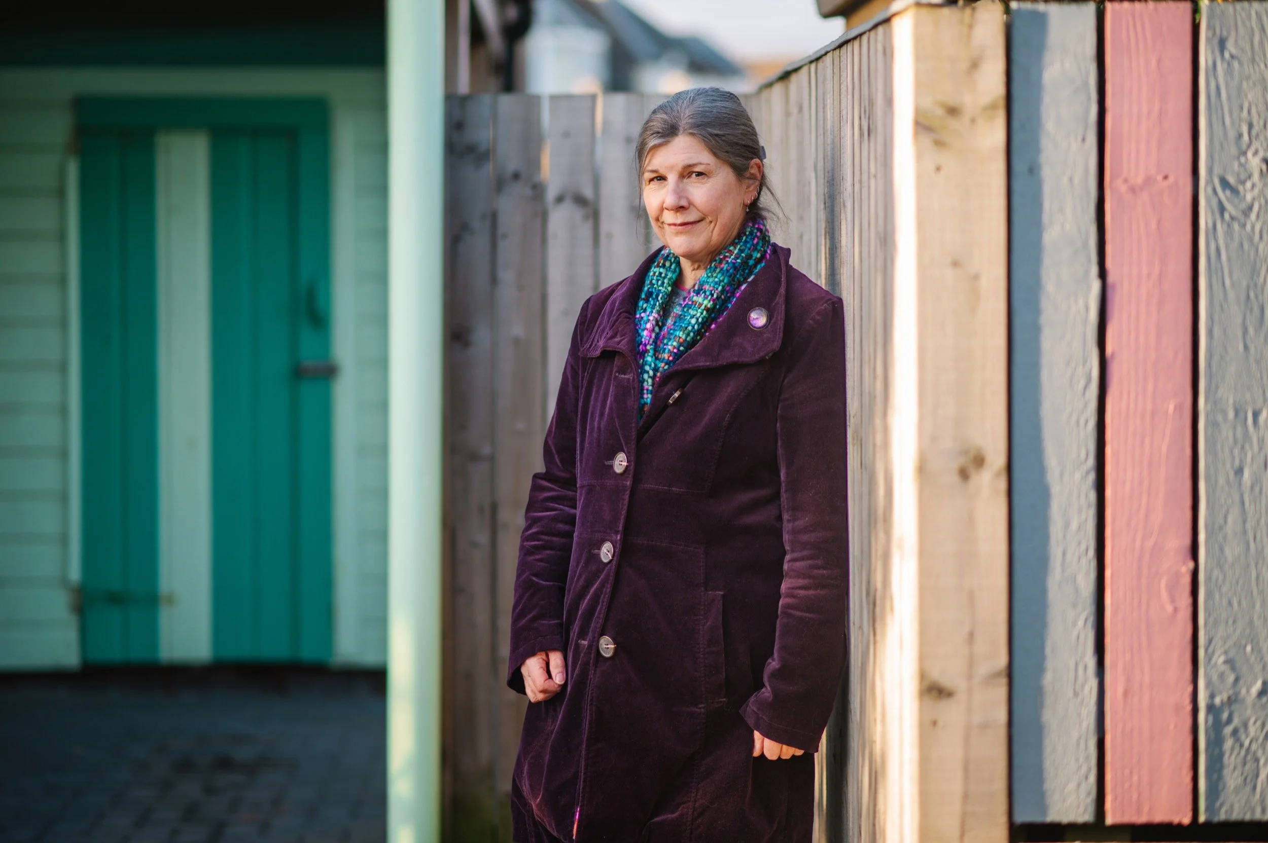 A woman in a purple coat standing against a wooden fence outdoors with a colourful building in the background.