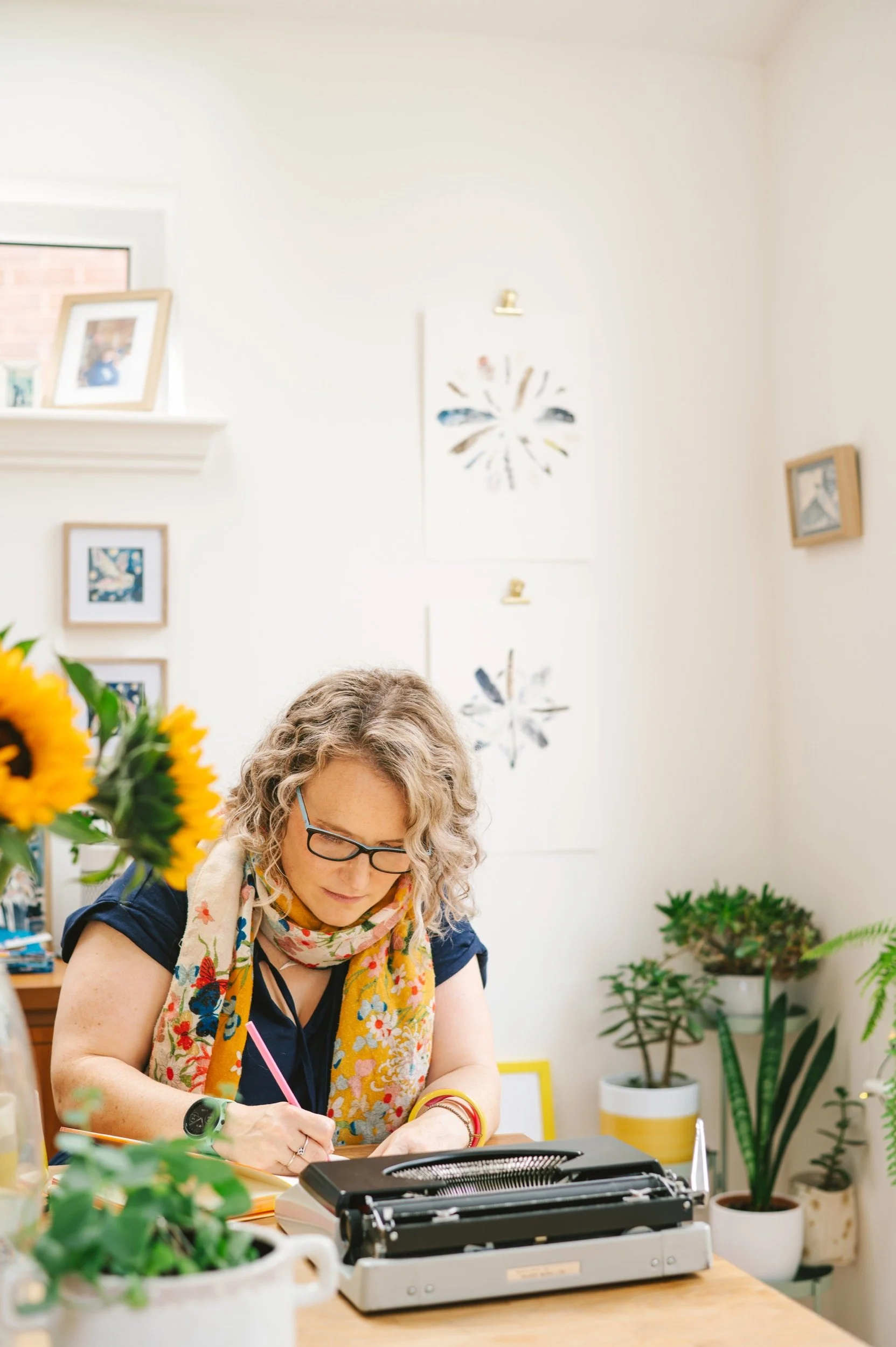 A woman with curly blonde hair, wearing glasses, a floral scarf, and a navy top, working at her desk with a typewriter, surrounded by potted plants and framed artwork on the walls.