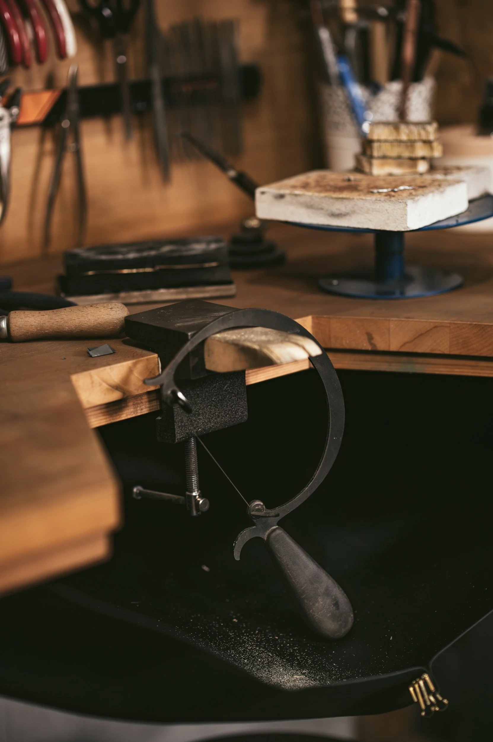 A jewellery bench with various tools, including a clamp, a file, and a small metal piece, with a cutting or polishing wheel and stacked wooden blocks in the background.