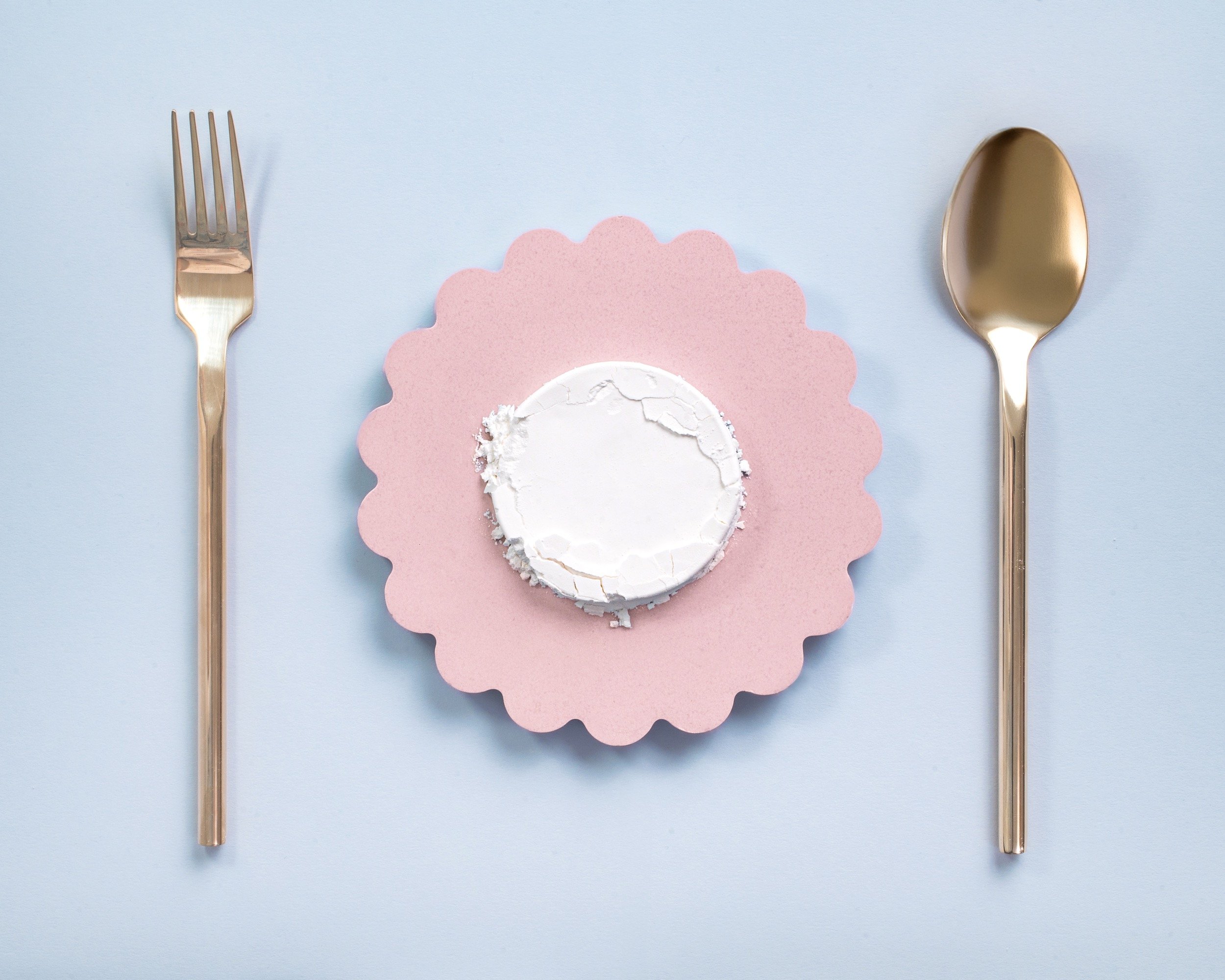 Cosmetic powder ingredient on a pink scalloped tray, with a gold fork on the left and a gold spoon on the right, all on a light blue background.