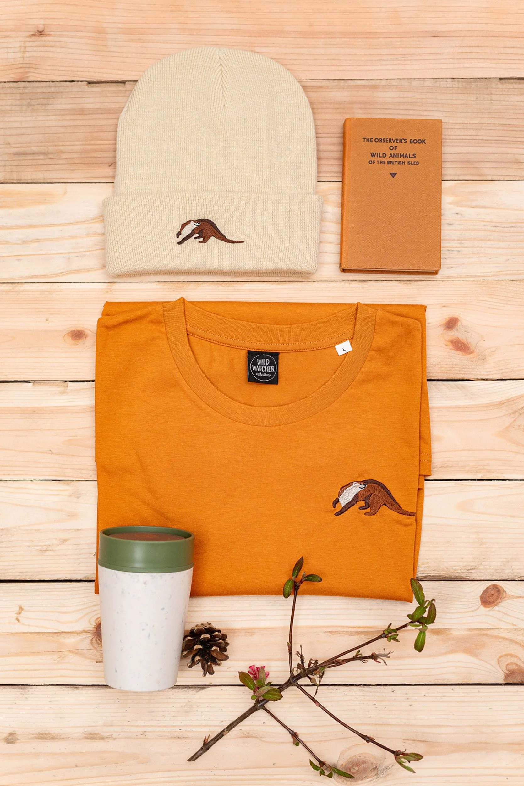 Items including a cream knit beanie with a small embroidered animal, an orange t-shirt with a matching embroidered animal, a small brown book titled 'The Observer's Book of Wild Animals of the British Isles', a white travel mug with a green lid, a pi