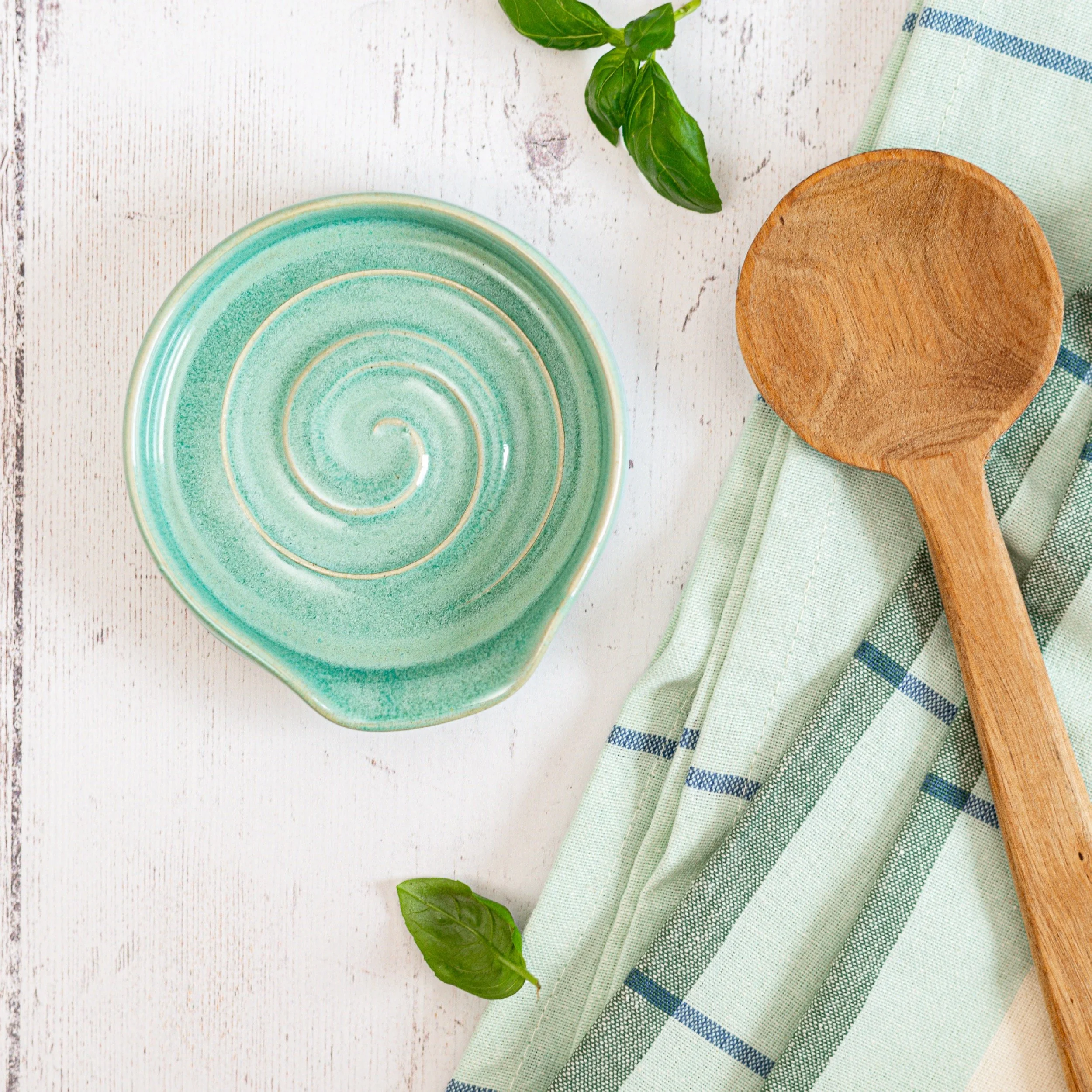 A green ceramic spiral-patterned spoon rest, a wooden spoon, a green and white striped napkin, and green basil leaves on a white wooden surface.