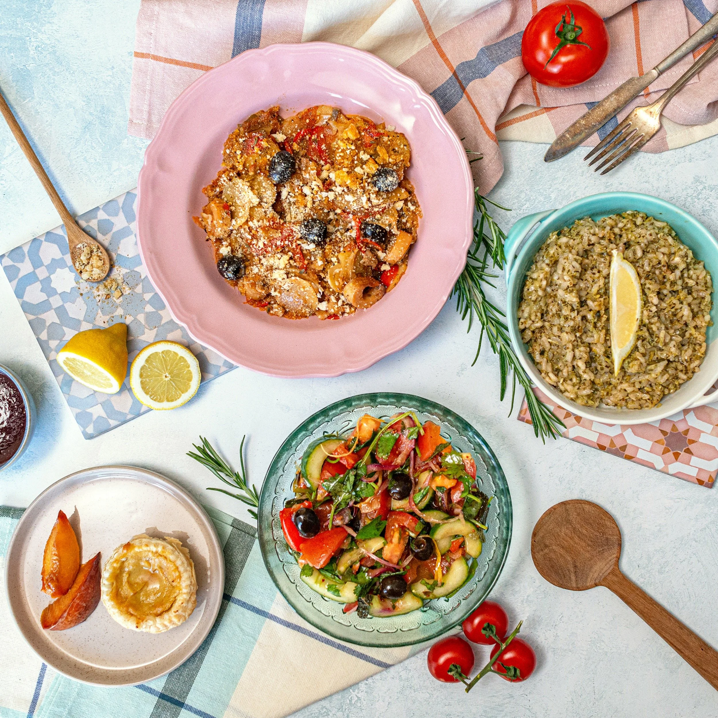 Assorted Mediterranean-style dishes on a table, including a vegetable casserole on a pink plate, a grain dish with lemon in a turquoise bowl, a fresh salad with cucumbers, tomatoes, and olives, set with lemons, tomatoes, and a wooden spoon.