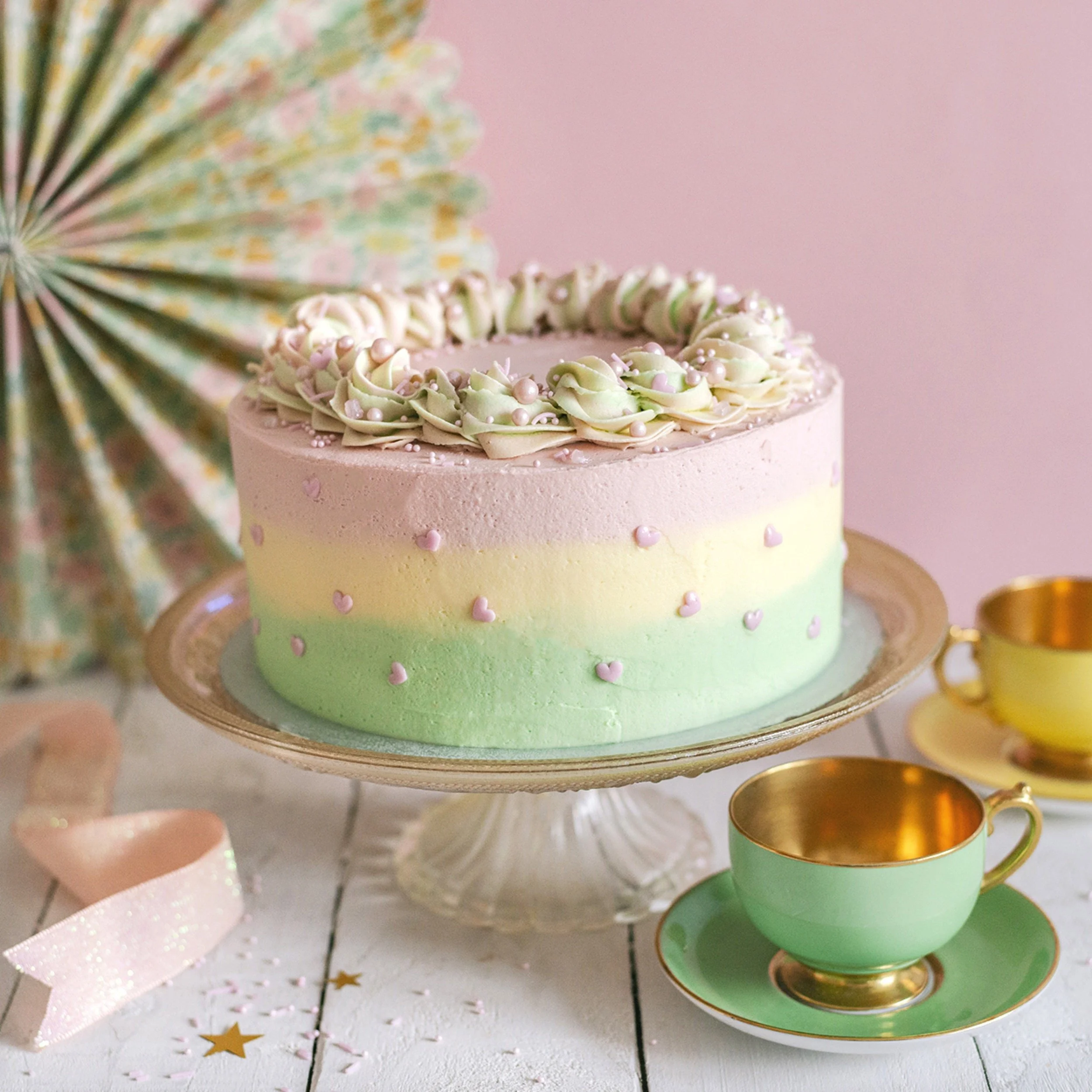 A pastel-colored cake with pink, yellow, and green layers, decorated with small heart sprinkles and piping on top, placed on a glass cake stand. There are gold and green teacups with matching saucers on a white wooden table, with a pink background an