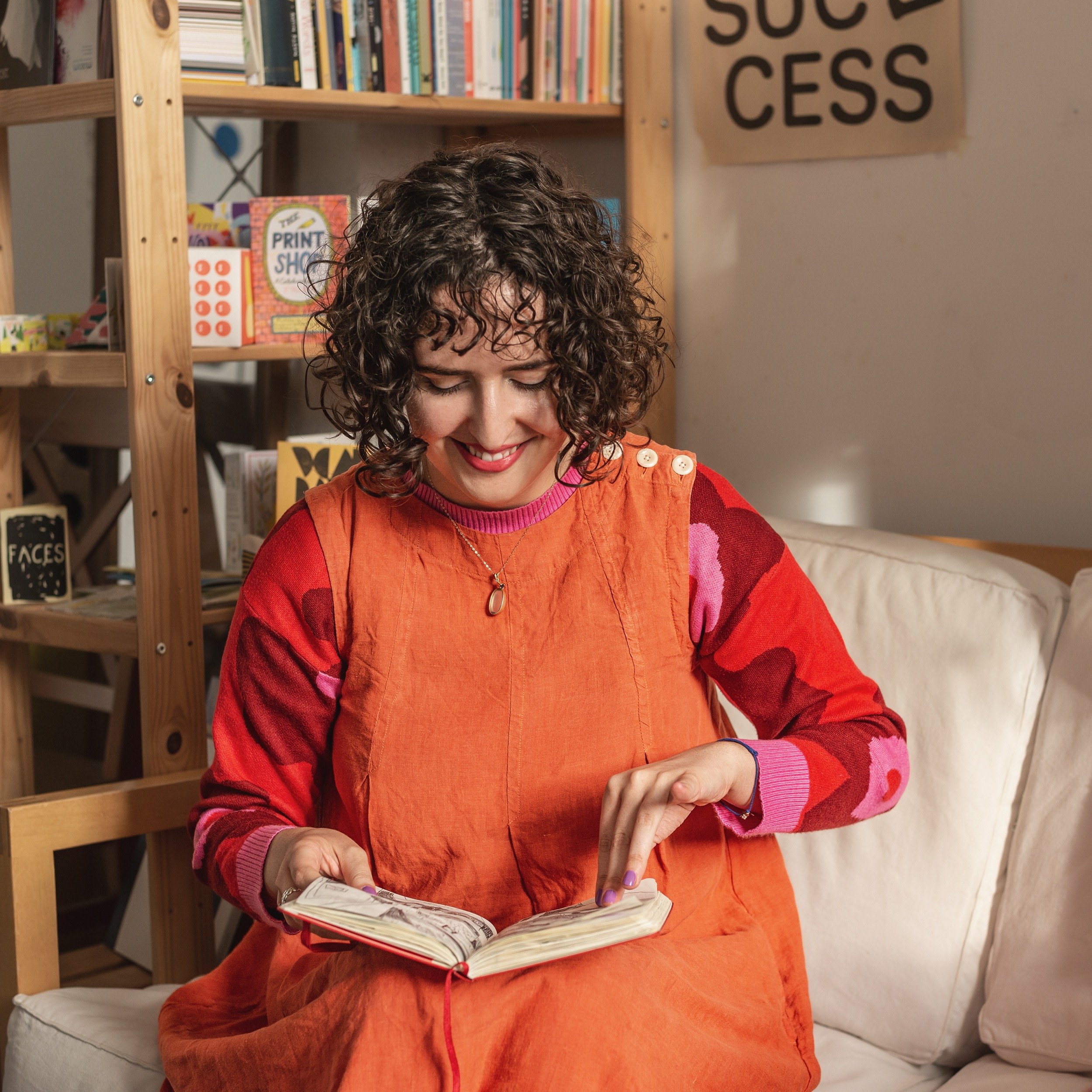A woman with curly hair wearing an orange dress and a colourful top with red, pink, and purple patterns, smiling and looking at a sketchbook in her hands while sitting on a beige couch in her studio.