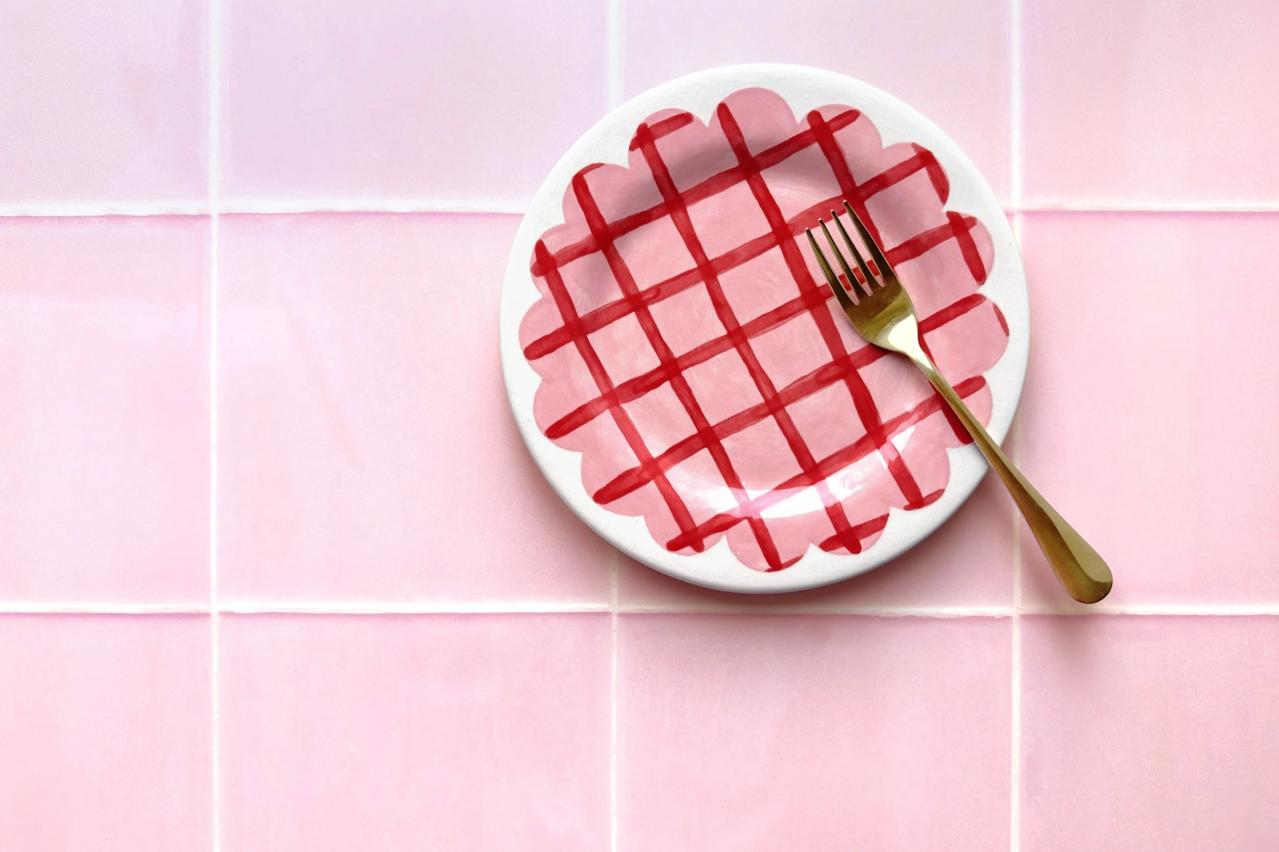 A white plate with a pink and red crisscross pattern, a gold fork resting on the right side, on a pink tiled surface.