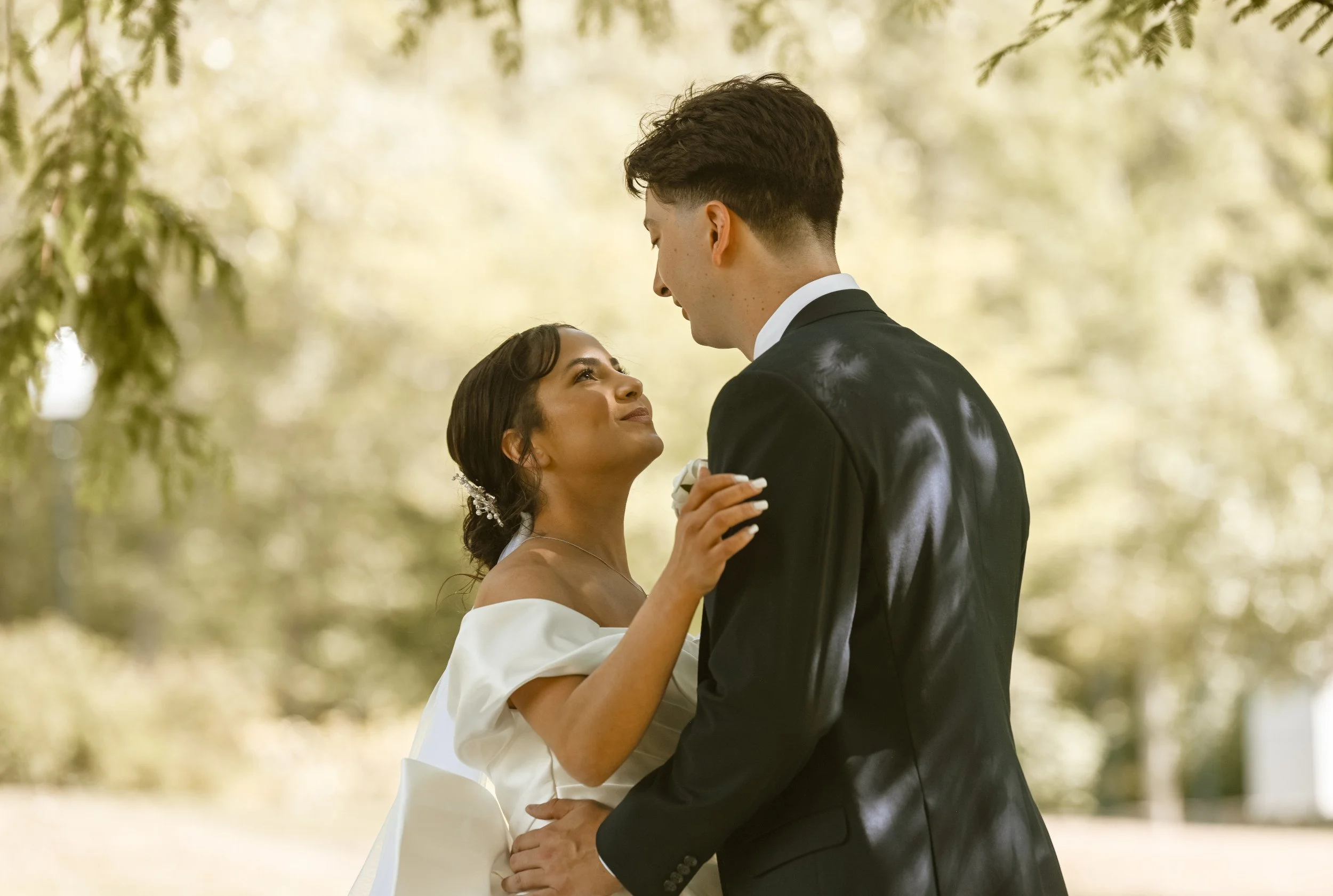 A bride and groom dancing outdoors, facing each other in a natural setting with trees in the background, during their wedding.