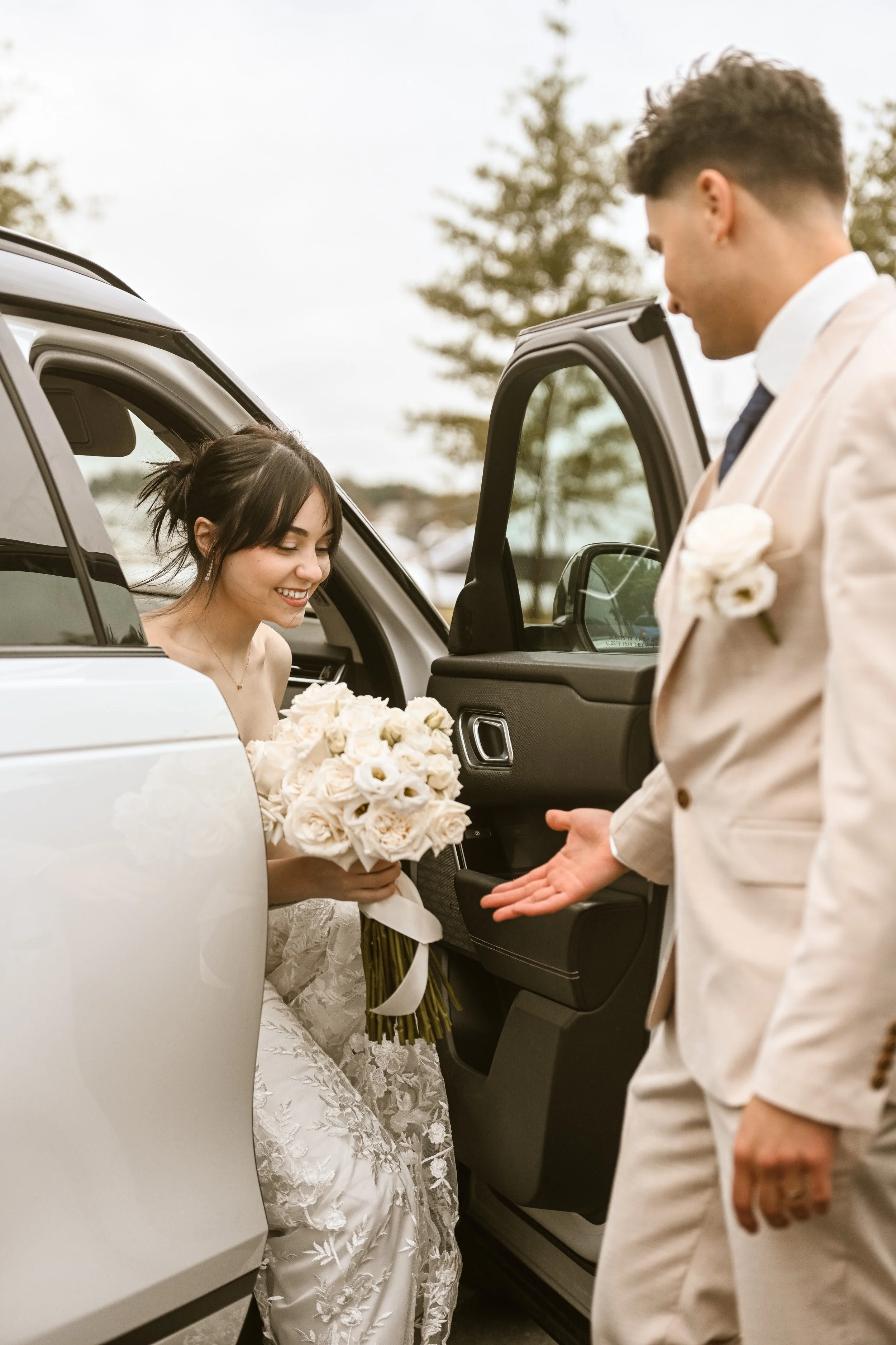 A bride smiling inside a white car, holding a bouquet of white roses, as a groom dressed in beige suit, white shirt, and black tie, extends his hand towards her during a wedding.