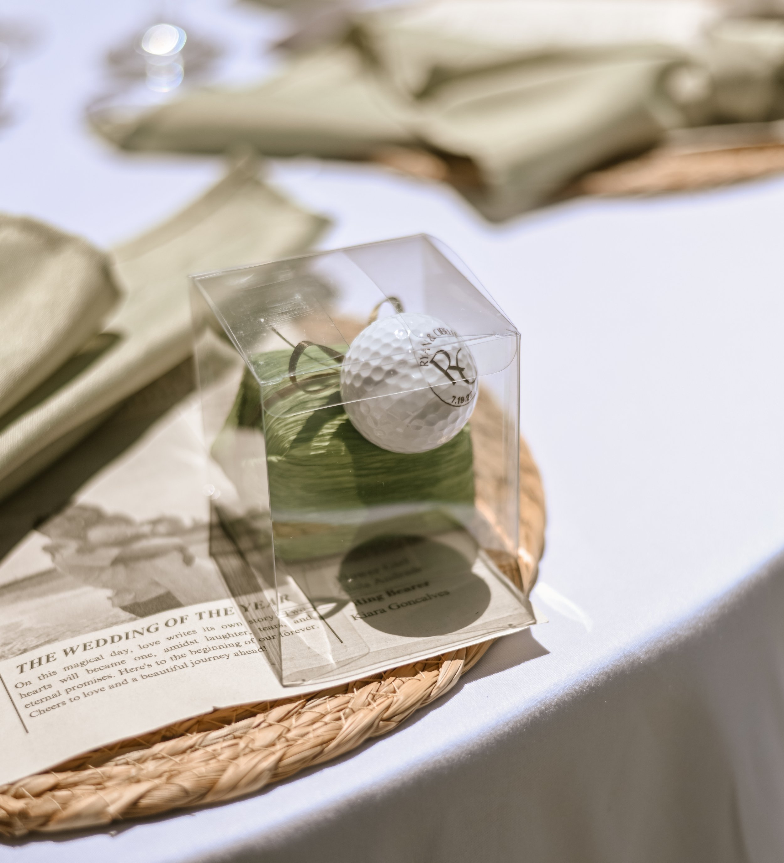 A small white golf ball-shaped item inside a clear acrylic box, resting on a green leaf, on top of a piece of paper with text, all on a woven tray.