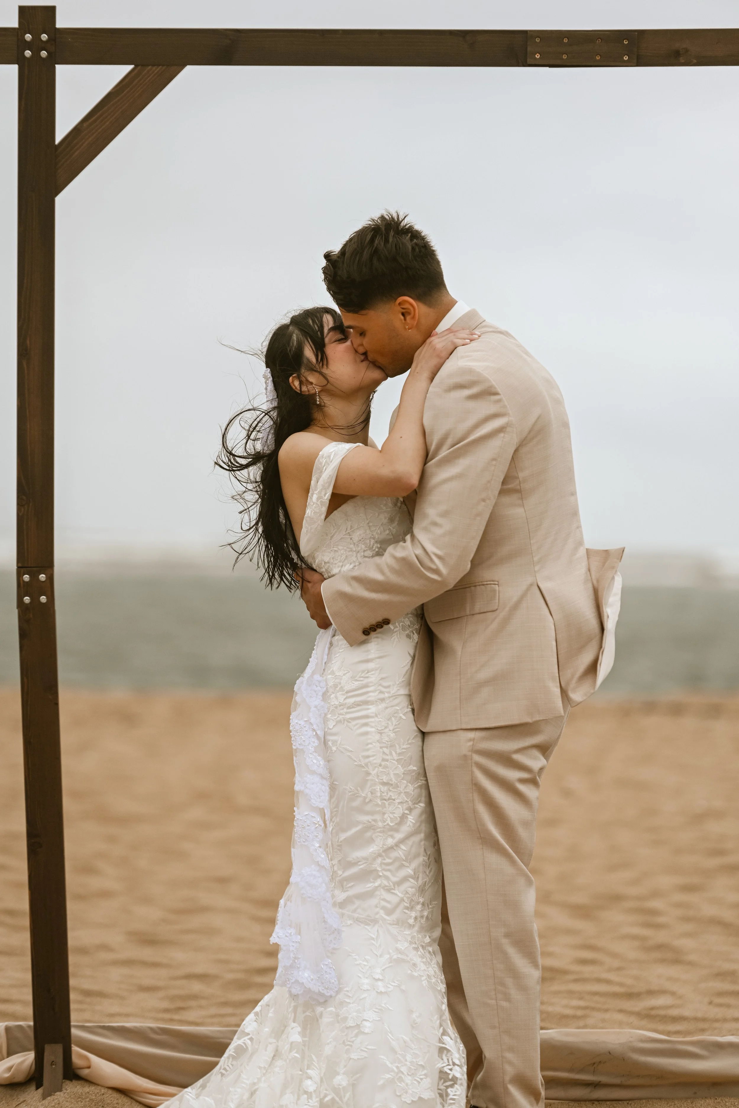 A bride and groom sharing a kiss during their wedding ceremony on the beach with the ocean in the background.