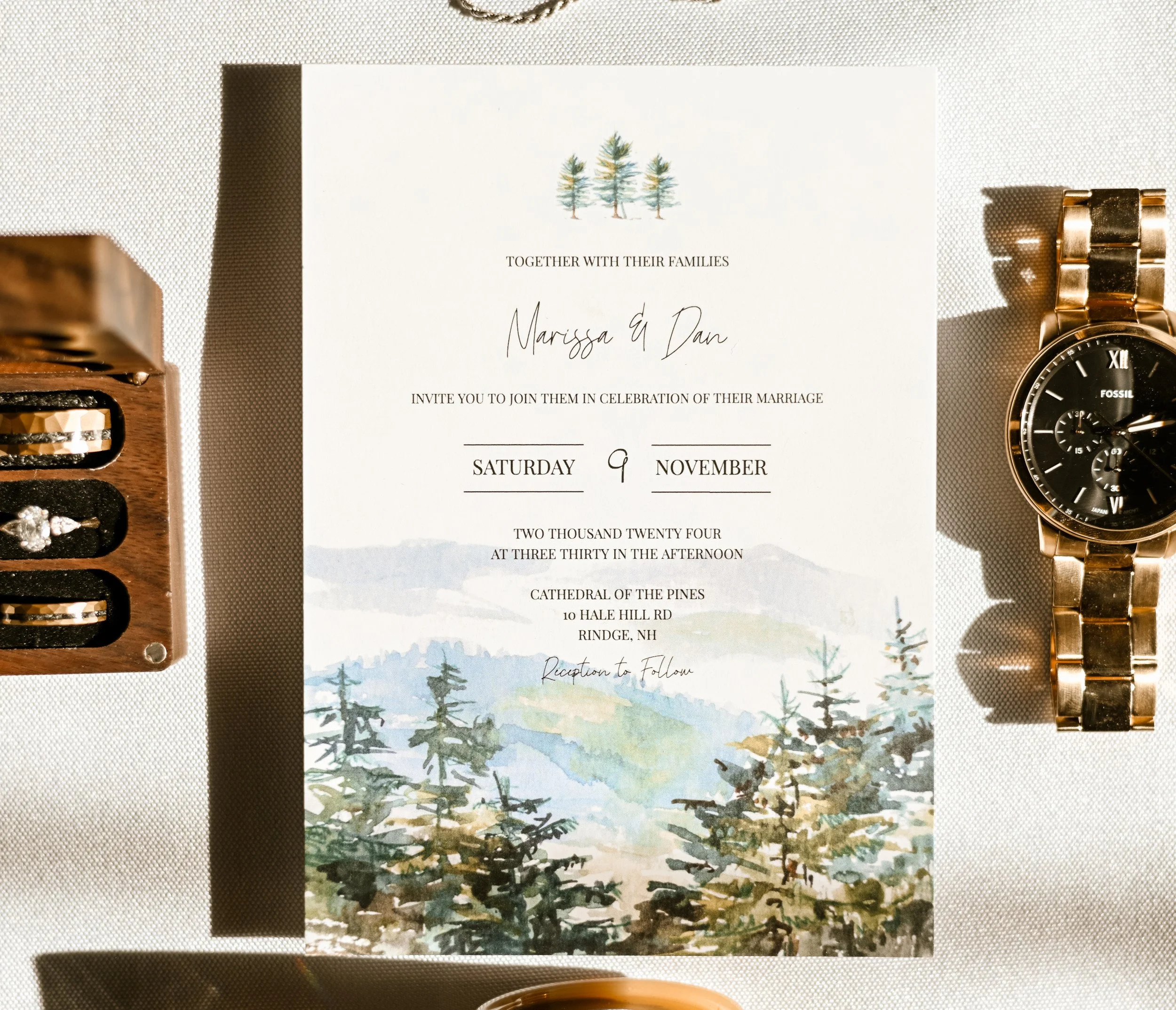 A wedding invitation with trees and mountains illustration, surrounded by a wooden box with jewelry on the left and a gold watch on the right. The invitation states the wedding date and location.