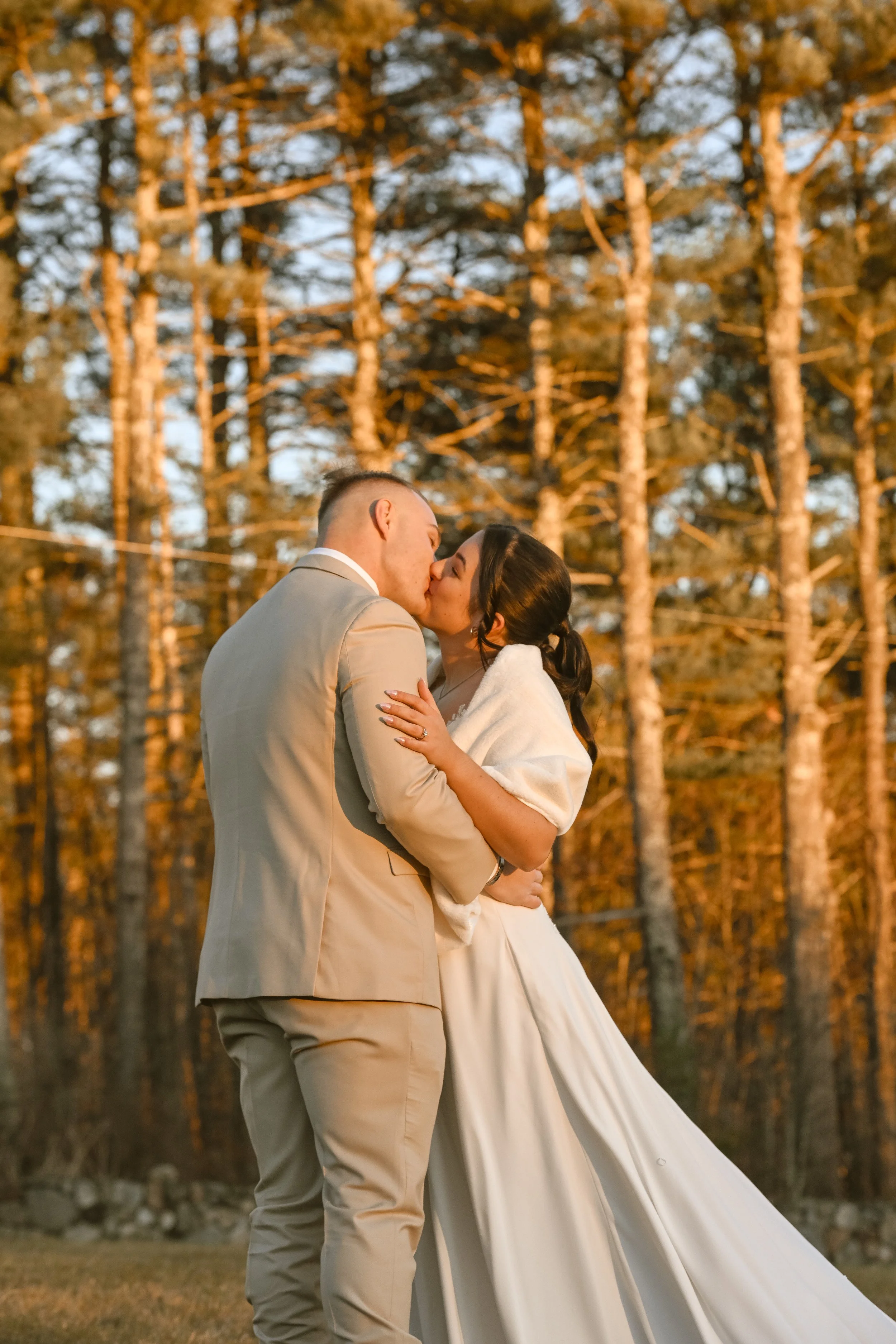 A couple on their wedding day sharing a kiss outdoors in front of a forest of tall trees during sunset.