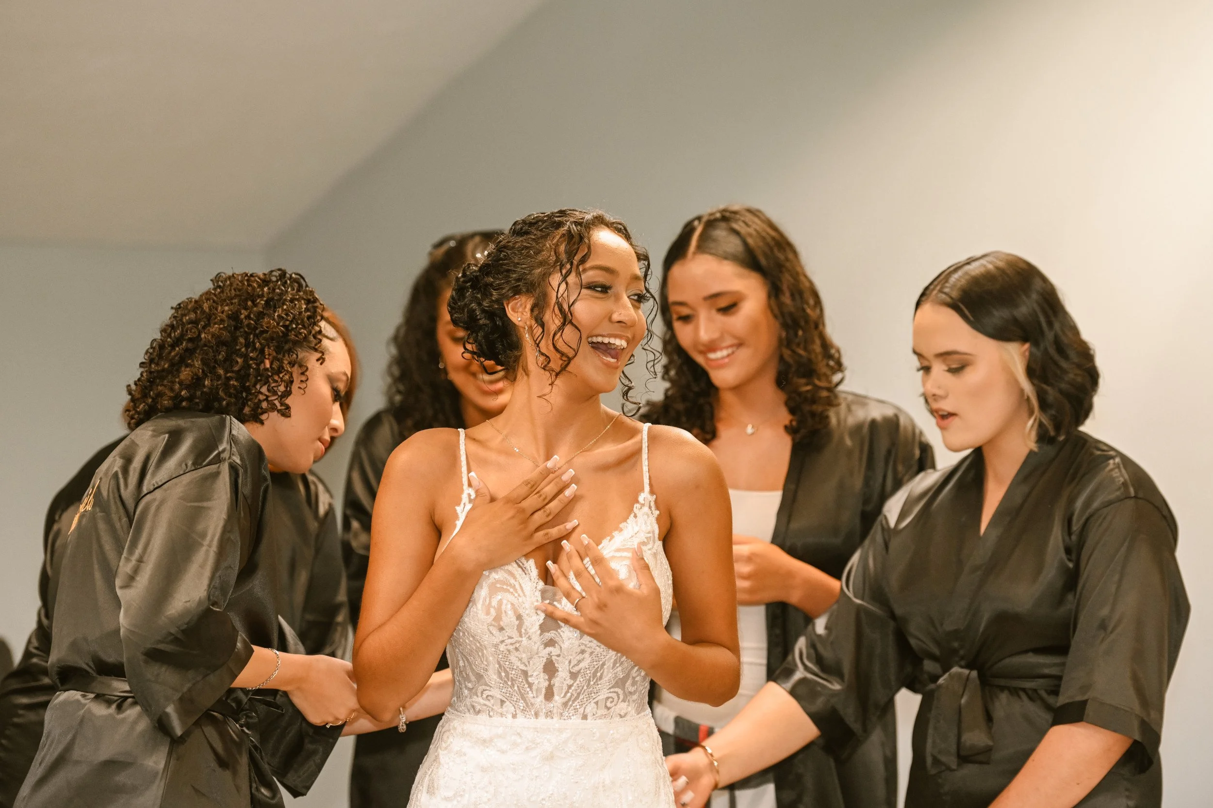 A bride is smiling with her hand on her chest as friends surround her, some wearing black robes, during a wedding preparation.
