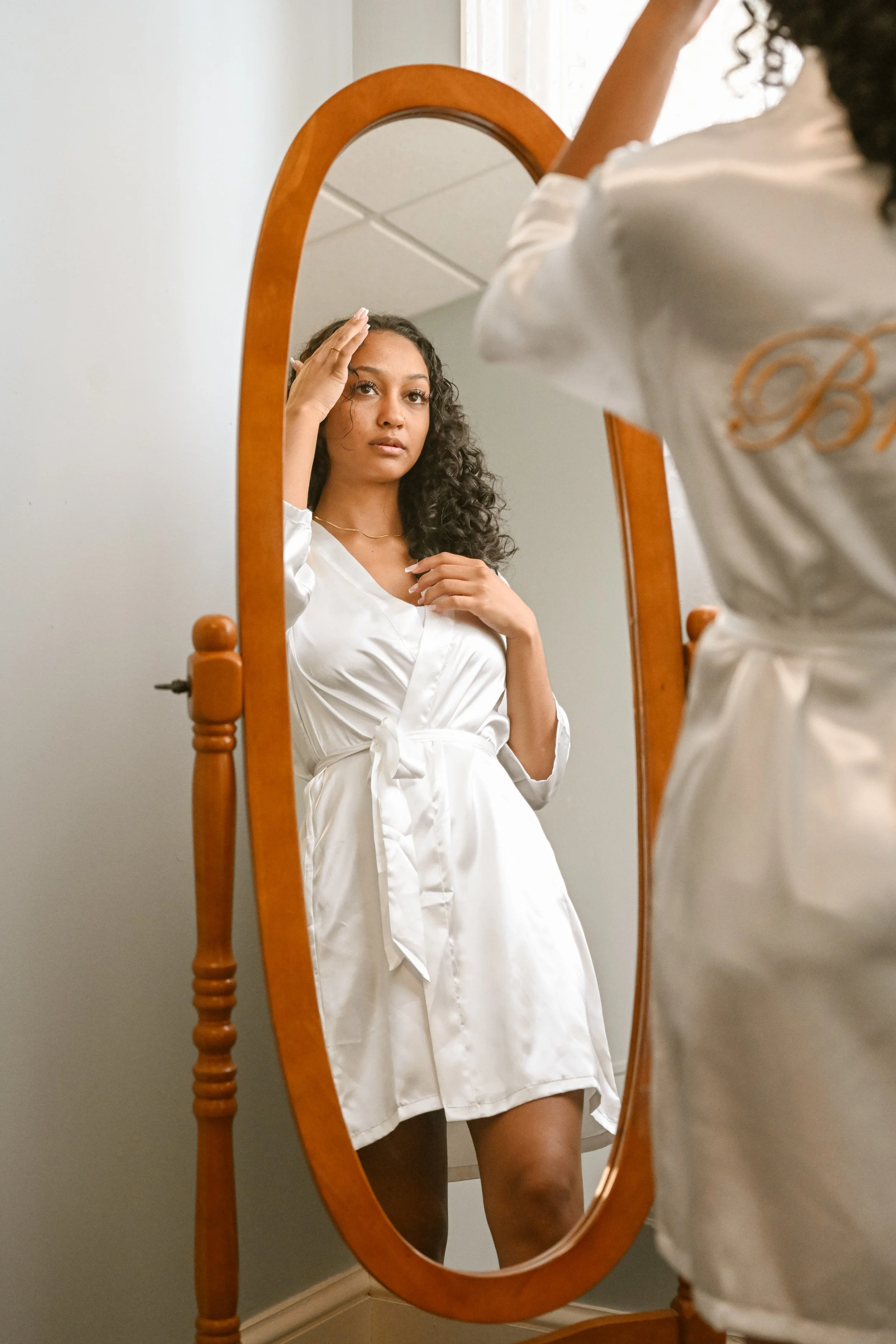 A woman with curly hair taking a selfie in a standing oval-shaped wooden mirror. She is wearing a white satin bathrobe with a tied waist, and is holding her hand to her forehead while looking at her reflection.